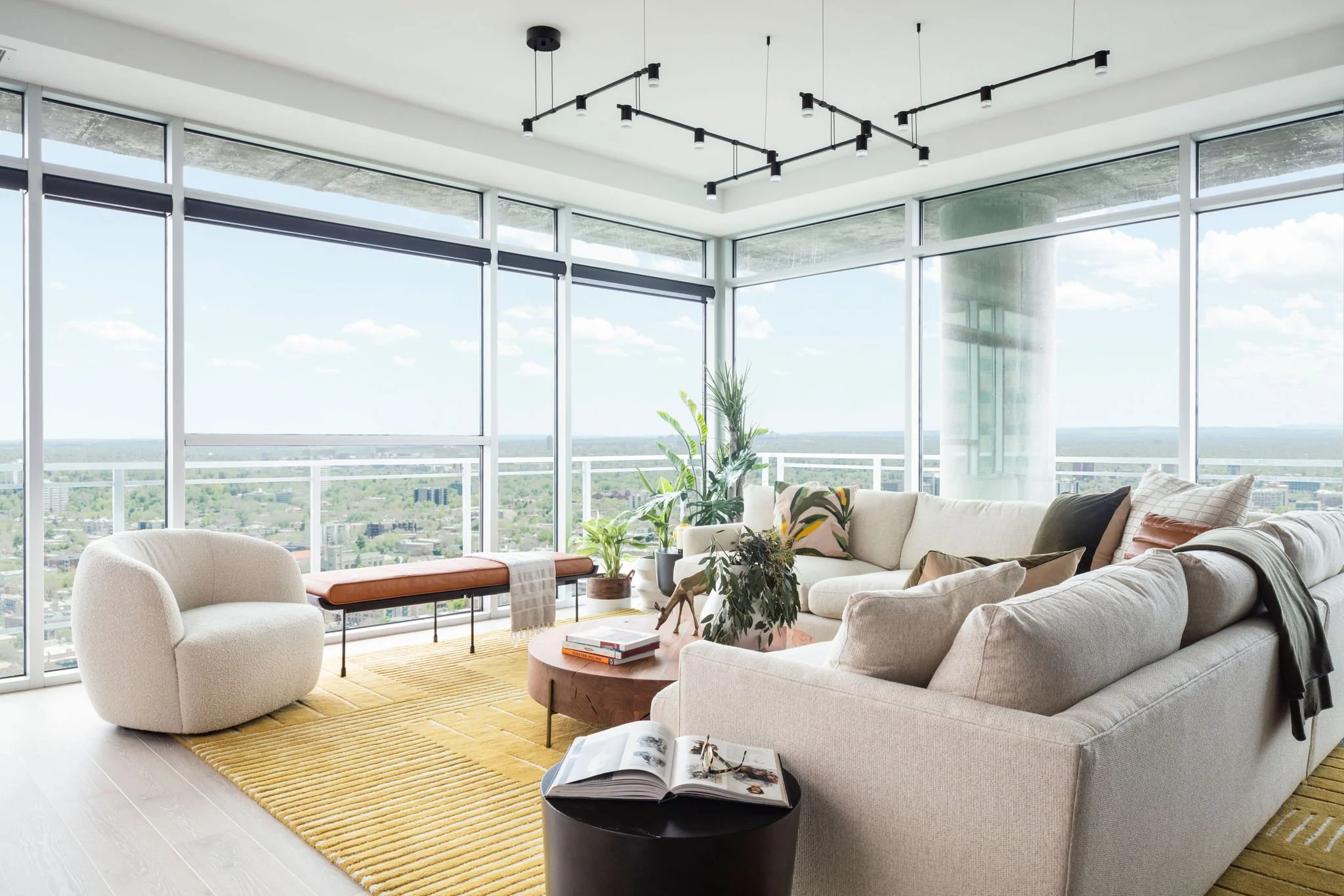 Modern living room with large floor-to-ceiling windows, white sectional sofa, white armchair, potted plants, wooden coffee table, and a yellow rug.