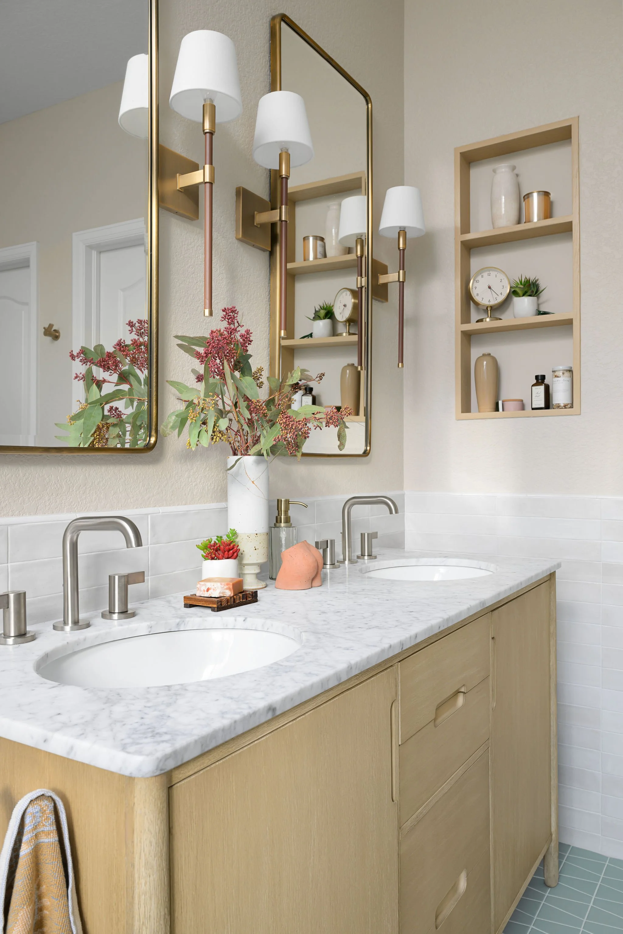 Bathroom vanity with marble countertop, two sinks, and modern faucets. Decor includes a large vase with pink and green foliage, smaller succulent, soap dispenser, and a pink decorative object. Two large mirrors with gold frames hang above the vanity. Open wall shelves hold vases, potted plants, clock, and small bottles. The bathroom has a light color scheme with textured beige walls and light green tiles.