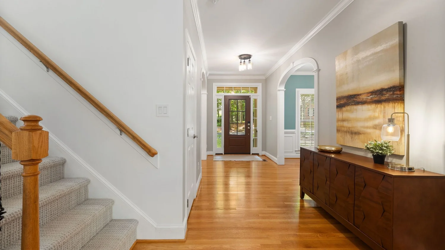 Front hallway of a renovated home with oak floors, a carpeted staircase with a wood newel post, an arched opening, and a wood credenza beneath a large abstract painting