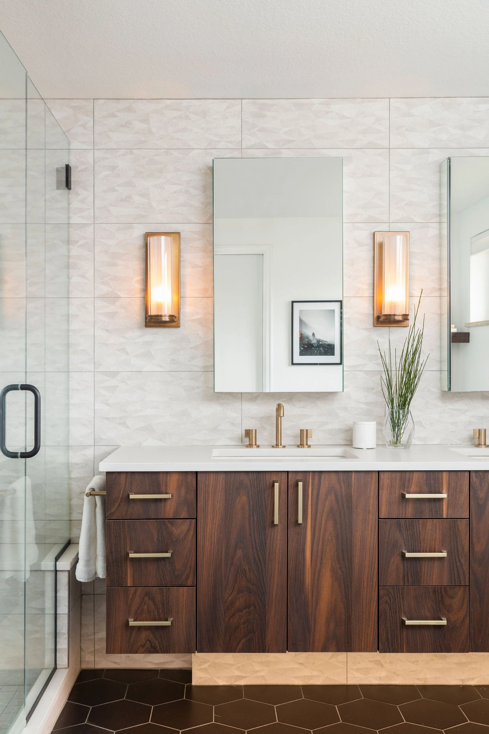 Modern bathroom vanity with wooden cabinet, white countertop, gold handles and fixtures, two mirrors with wall sconces, a vase with green plants, and a shower with glass door on the left.