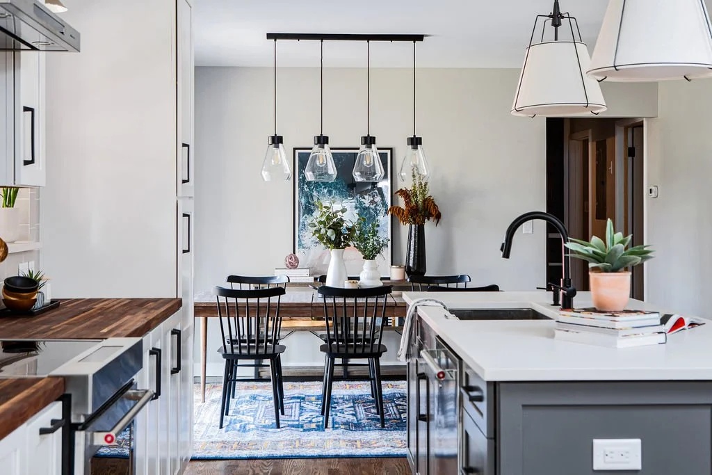 Modern kitchen with black chairs at a wooden dining table, hanging pendant lights, and decorative plants on the white countertop and table.