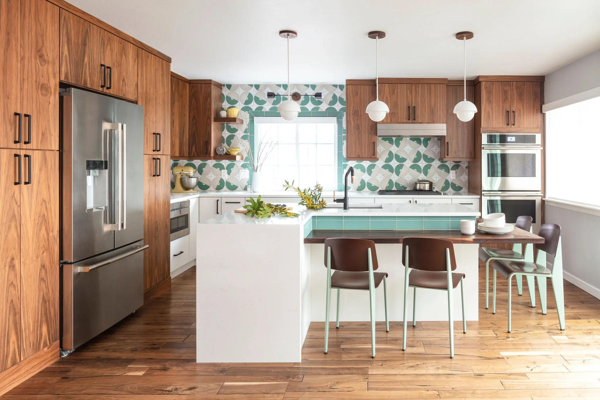 Modern kitchen with wood cabinets, white countertops, and a patterned backsplash. There is a stainless steel refrigerator, built-in oven, and a kitchen island with green tile accents. Four brown chairs are arranged around a small dining table. Pendant lights hang from the ceiling, and a window provides natural light.