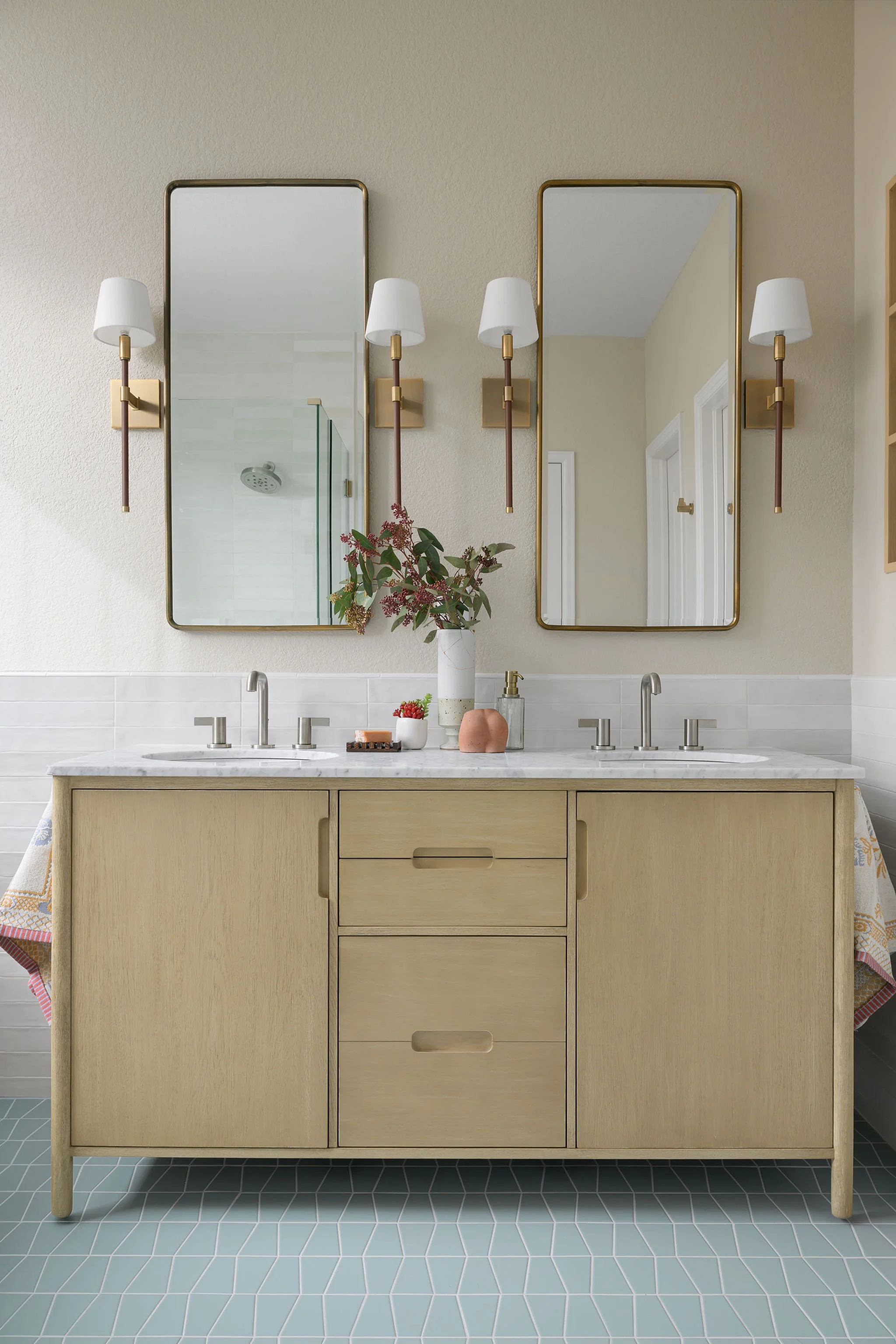 A modern bathroom vanity with double sinks, two rectangular mirrors, and two wall sconces with white shades. There are decorative objects on the countertop, including a vase with flowers, a soap dispenser, and small containers. The vanity is against a light-colored textured wall, and the floor has teal hexagonal tiles.