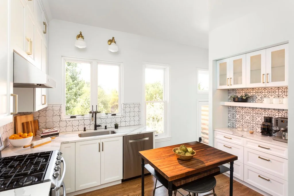 Bright, modern kitchen with white cabinets, black hardware, a wood countertop on the island, and geometric patterned tile backsplash. Large windows with greenery outside, black faucet, and small appliances on the counter.