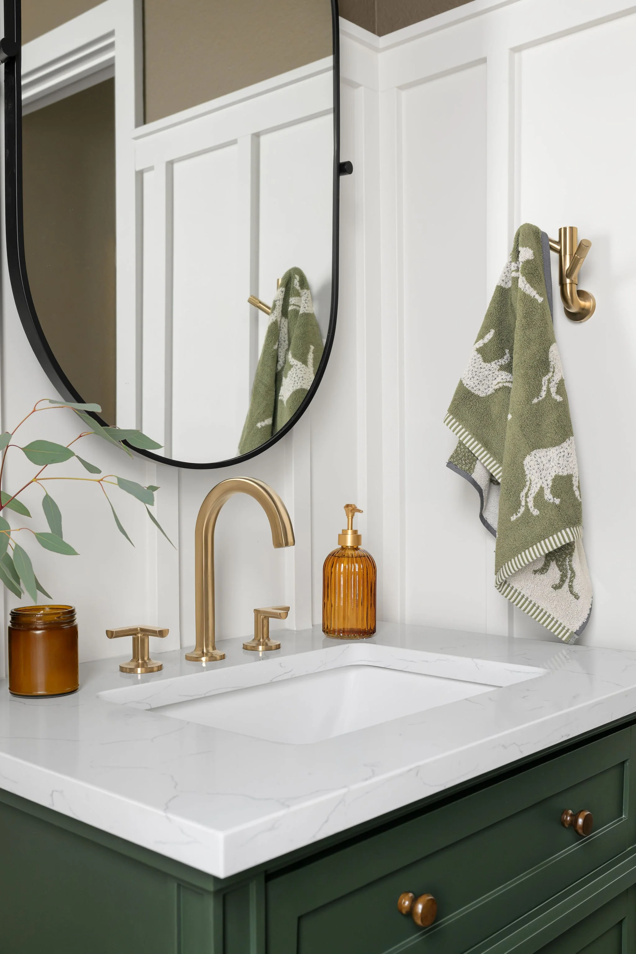 Bathroom vanity with gold fixtures, green cabinets, white marble countertop, and a mirror. A hand soap dispenser and a small jar are on the counter. Two green and white towels with animal designs hang on gold hooks, reflected in the mirror.