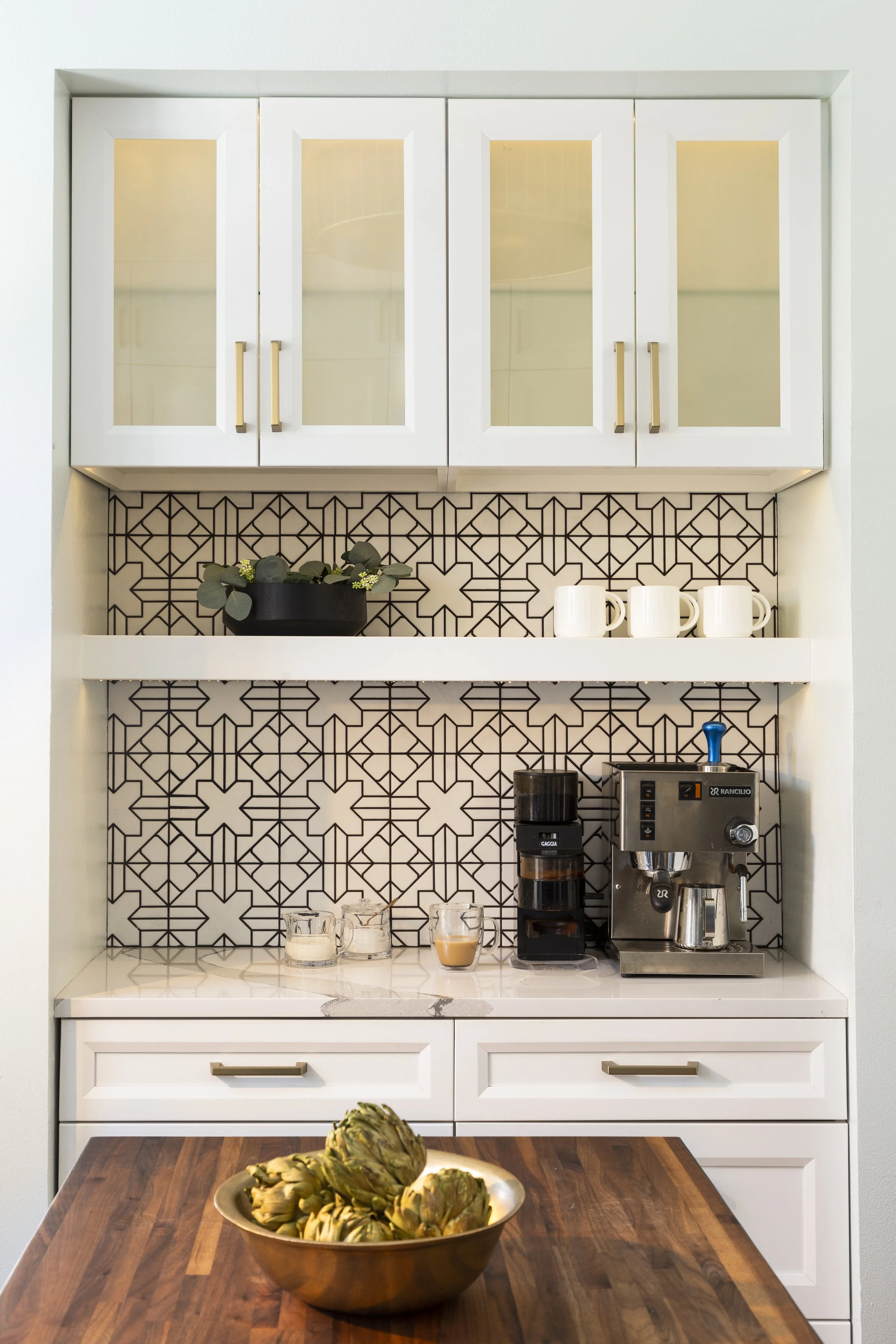 A kitchen nook with white cabinets with glass doors, a decorative tile backsplash, a coffee maker, a coffee grinder, three white mugs, a bowl of artichokes, and a countertop with three glass cups and a small cup of coffee.