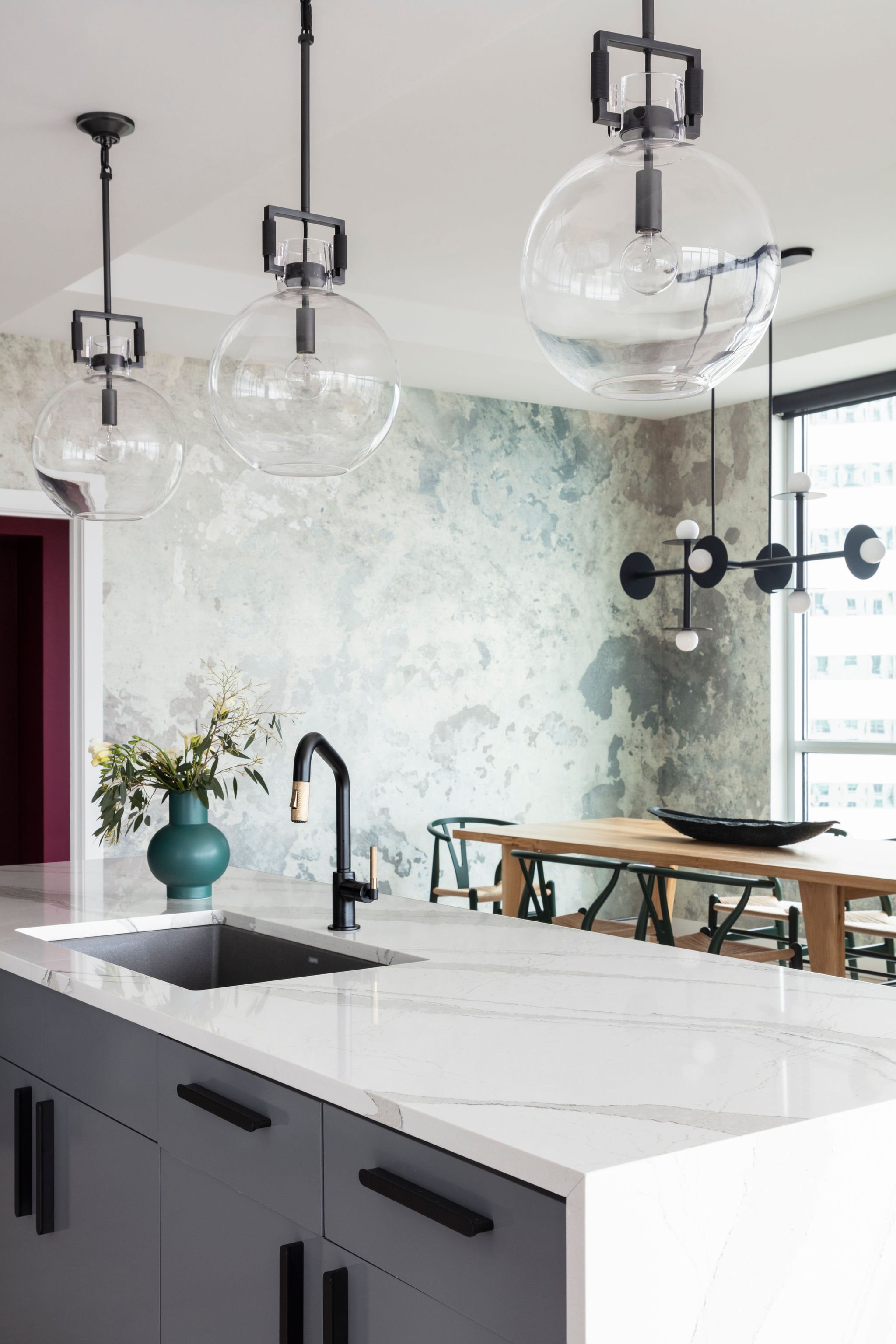 Modern kitchen with a white marble island, black faucet, vase with greenery, three glass globe pendant lights, and a contemporary black and white chandelier, with a dining table and chairs in the background near large windows.