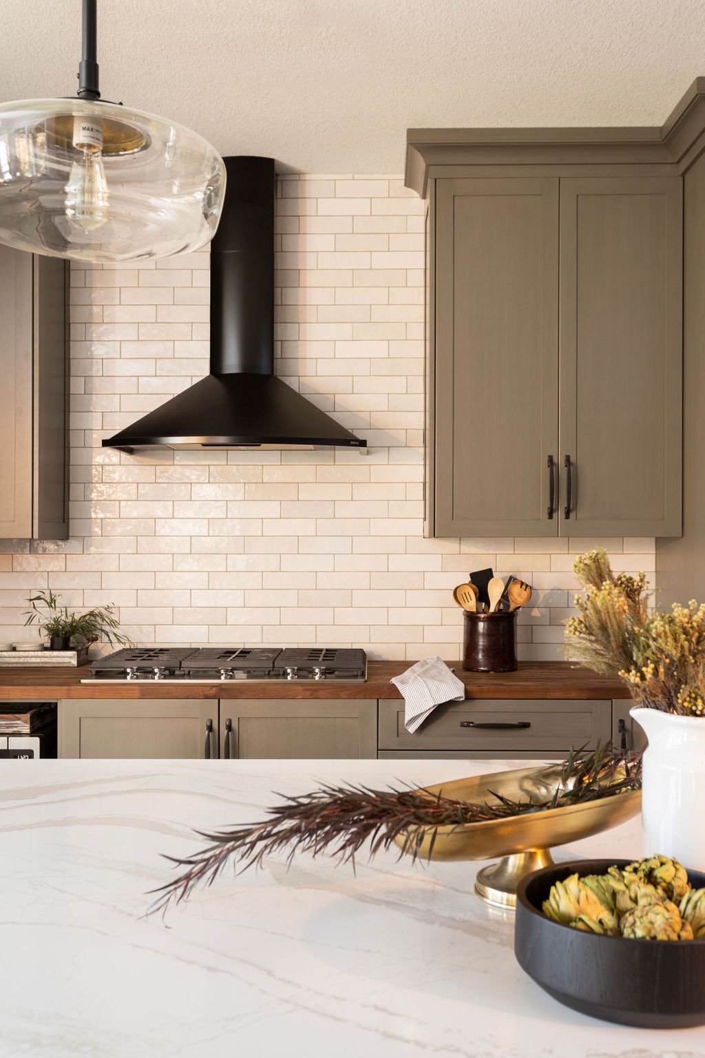 Modern kitchen with light gray cabinets, a white brick backsplash, a black range hood, and a wooden countertop. Decor includes a vase with dried flowers, a bowl of artichokes, and some kitchen utensils in a holder.