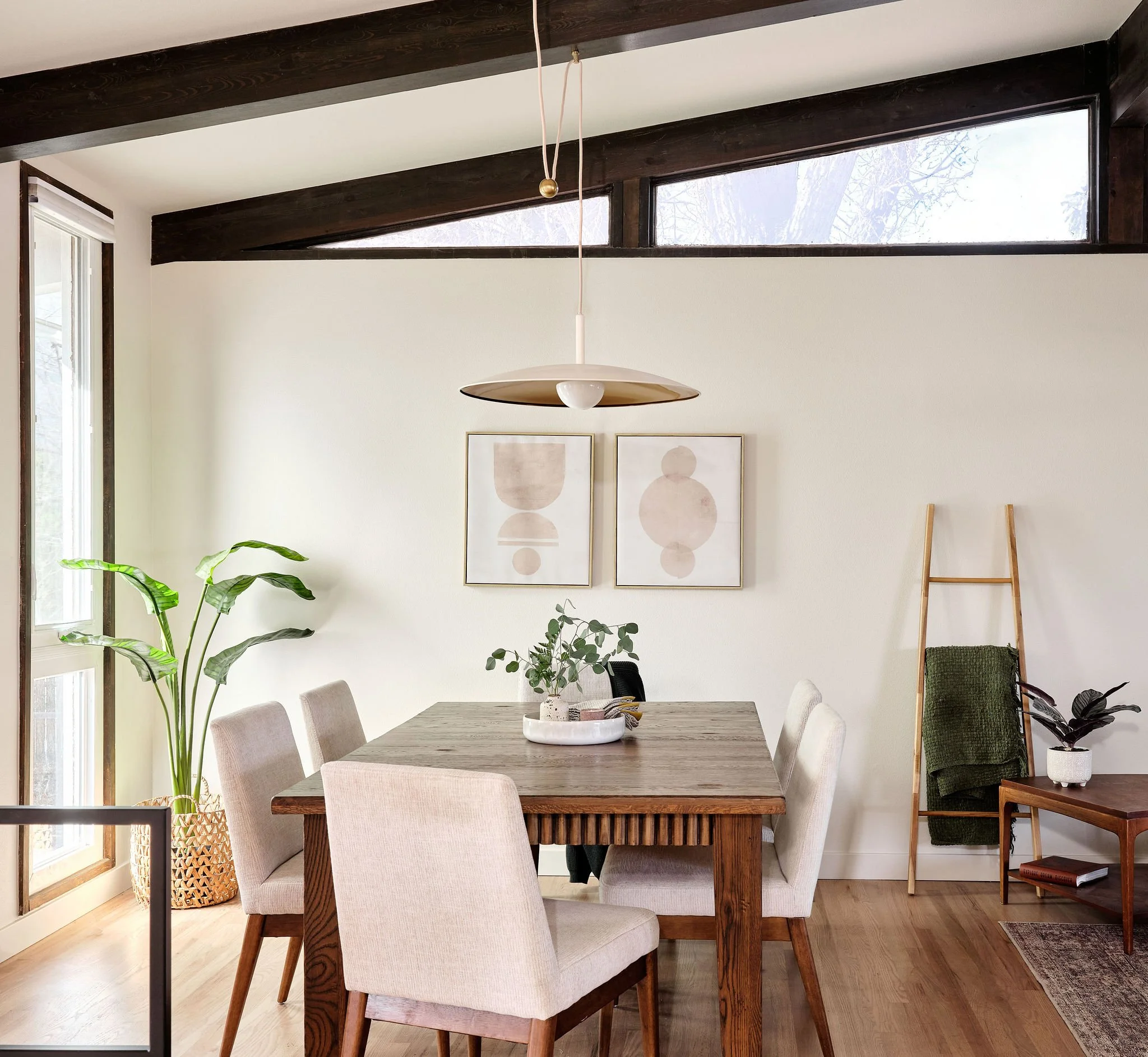 A dining room with a wooden table and six beige upholstered chairs, a large green plant in a woven basket, and wall art with abstract drawings. A pendant light hangs above the table, and a wooden ladder with a dark green towel is against the wall. Large windows let in natural light.