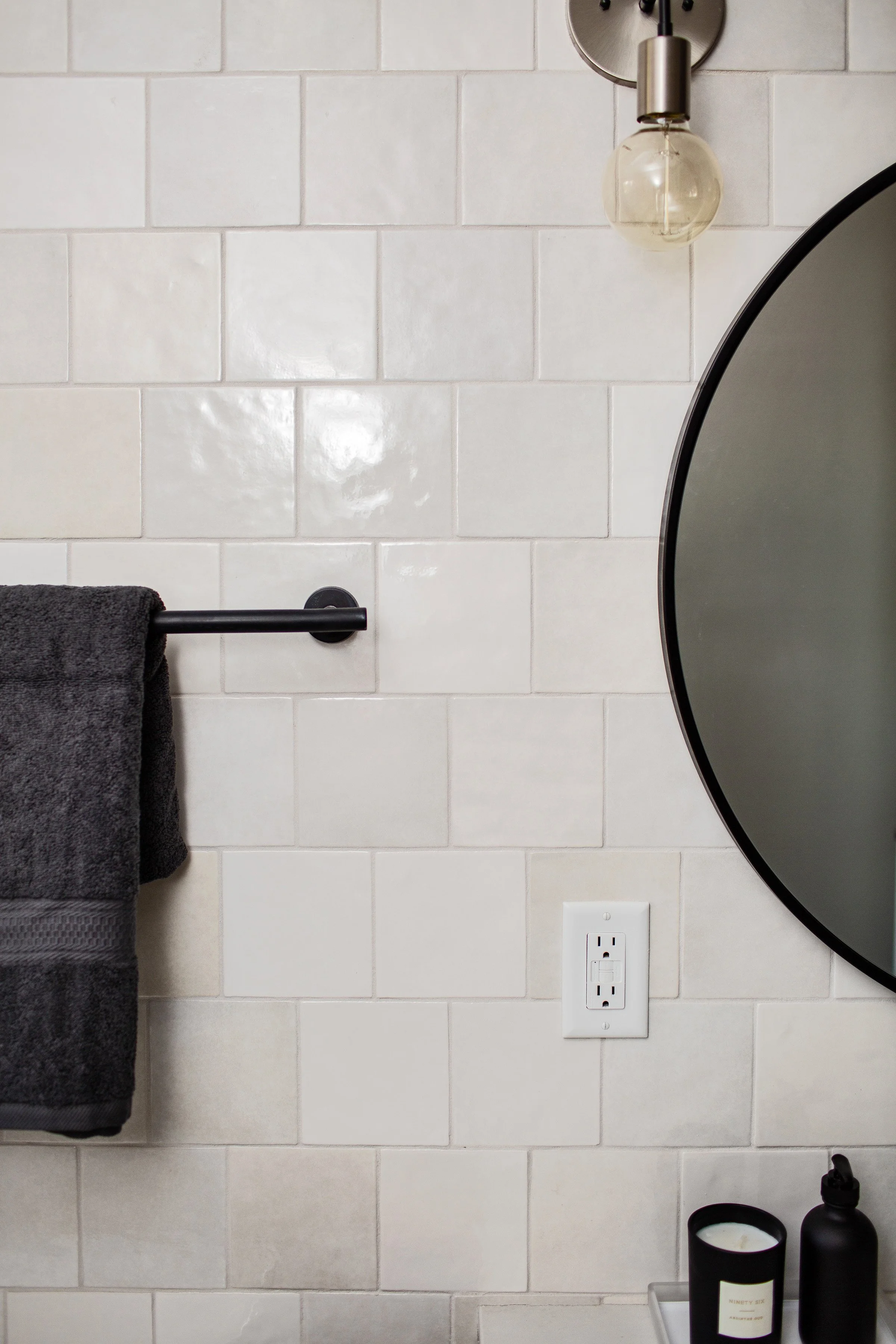 Bathroom with a towel rack holding a dark gray towel, a round mirror, a wall-mounted light fixture with a visible light bulb, a white electrical outlet, and a soap dispenser with a candle on a tray.