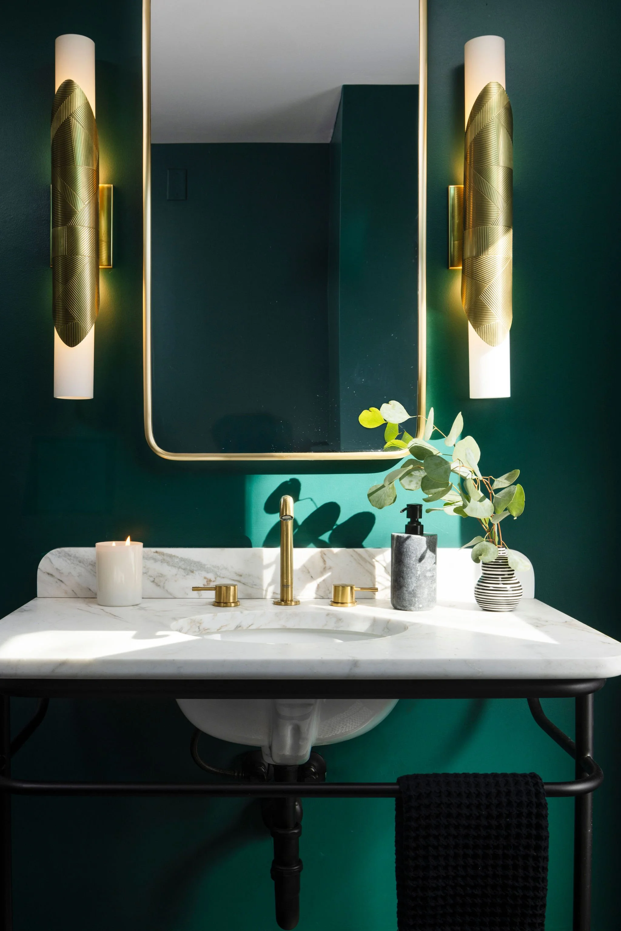 Bathroom with a white marble sink, gold fixtures, a framed mirror, and wall-mounted gold and white lights, with a green wall, a candle, a soap dispenser, and a vase with eucalyptus leaves.