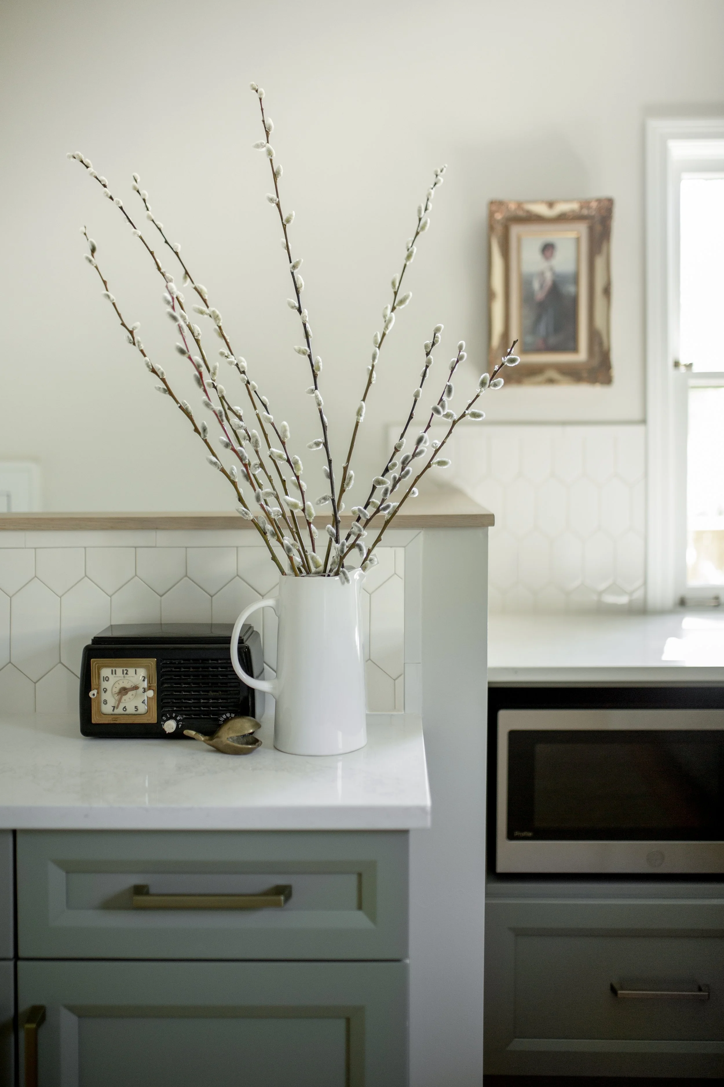 A white pitcher with pussy willow branches on a kitchen counter, with a vintage clock and a small bird figurine nearby, and a framed painting on the wall in the background.
