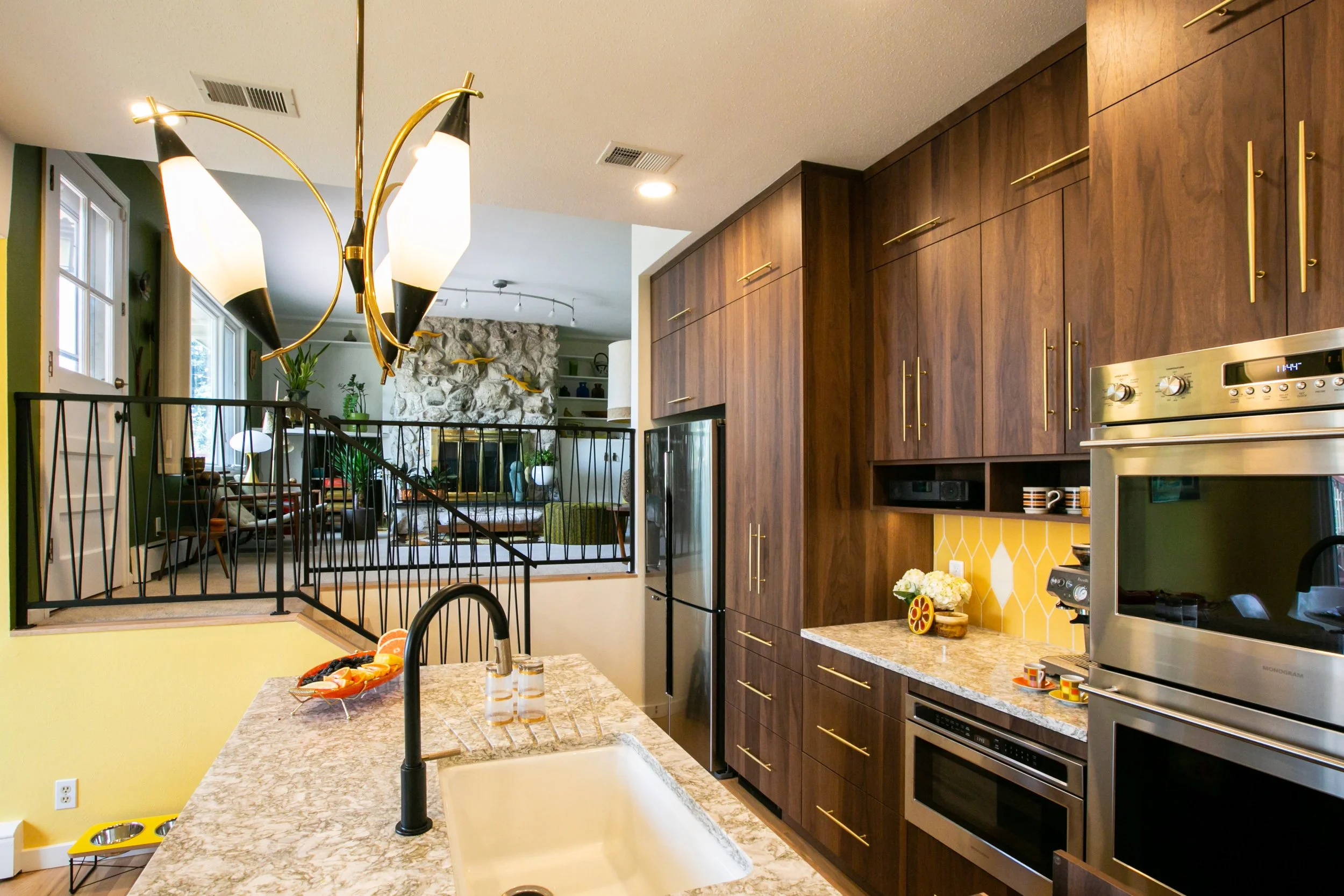 Modern kitchen with dark wood cabinets, stainless steel appliances, yellow tile backsplash, granite countertops, black faucet, and a breakfast bar with fruit bowl, with a view into a living room with stone fireplace and seating area.