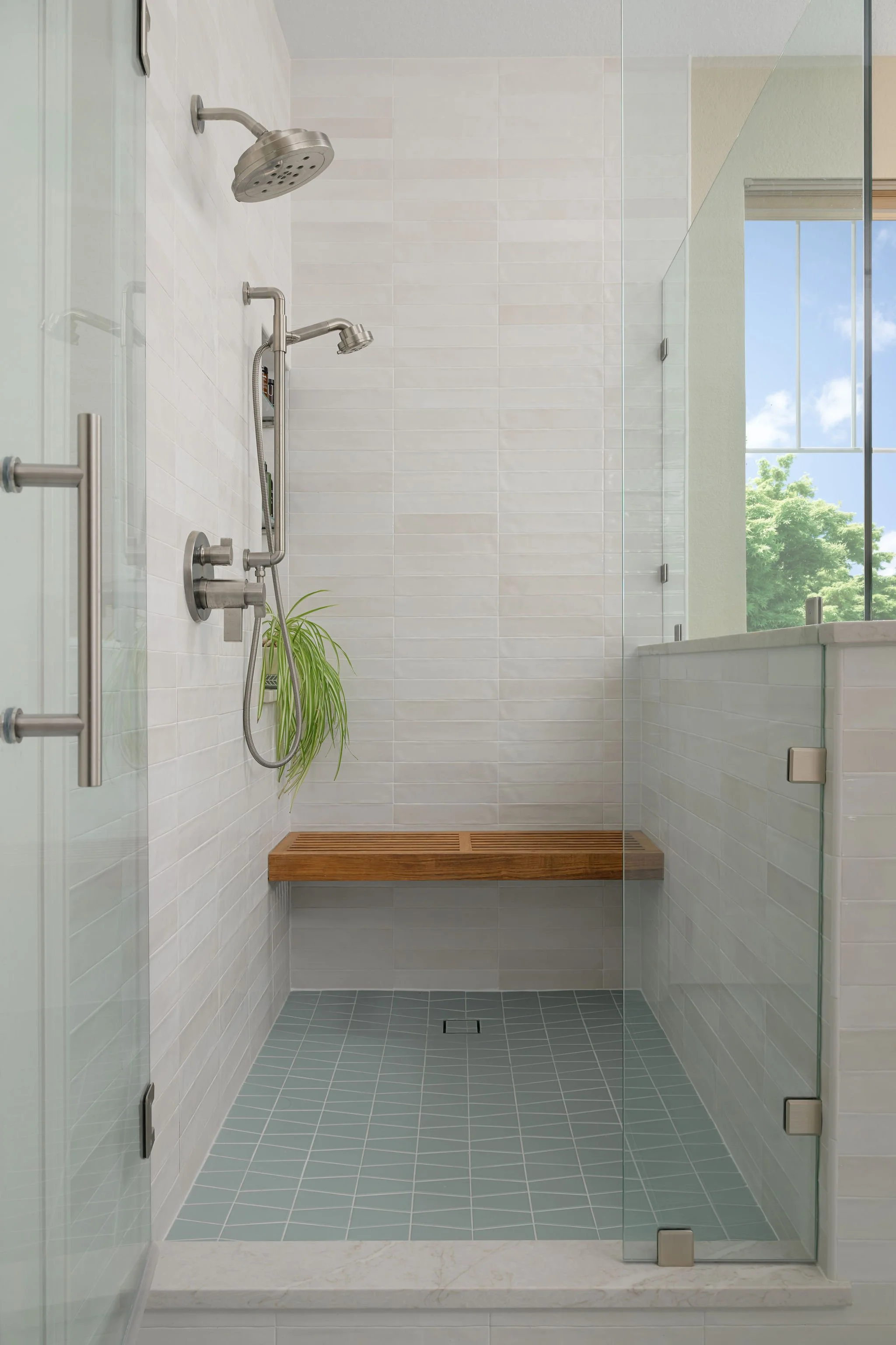 Modern glass-enclosed walk-in shower with two shower heads, built-in bench, white tiled walls, gray tiled floor, and large window showing green trees outside.