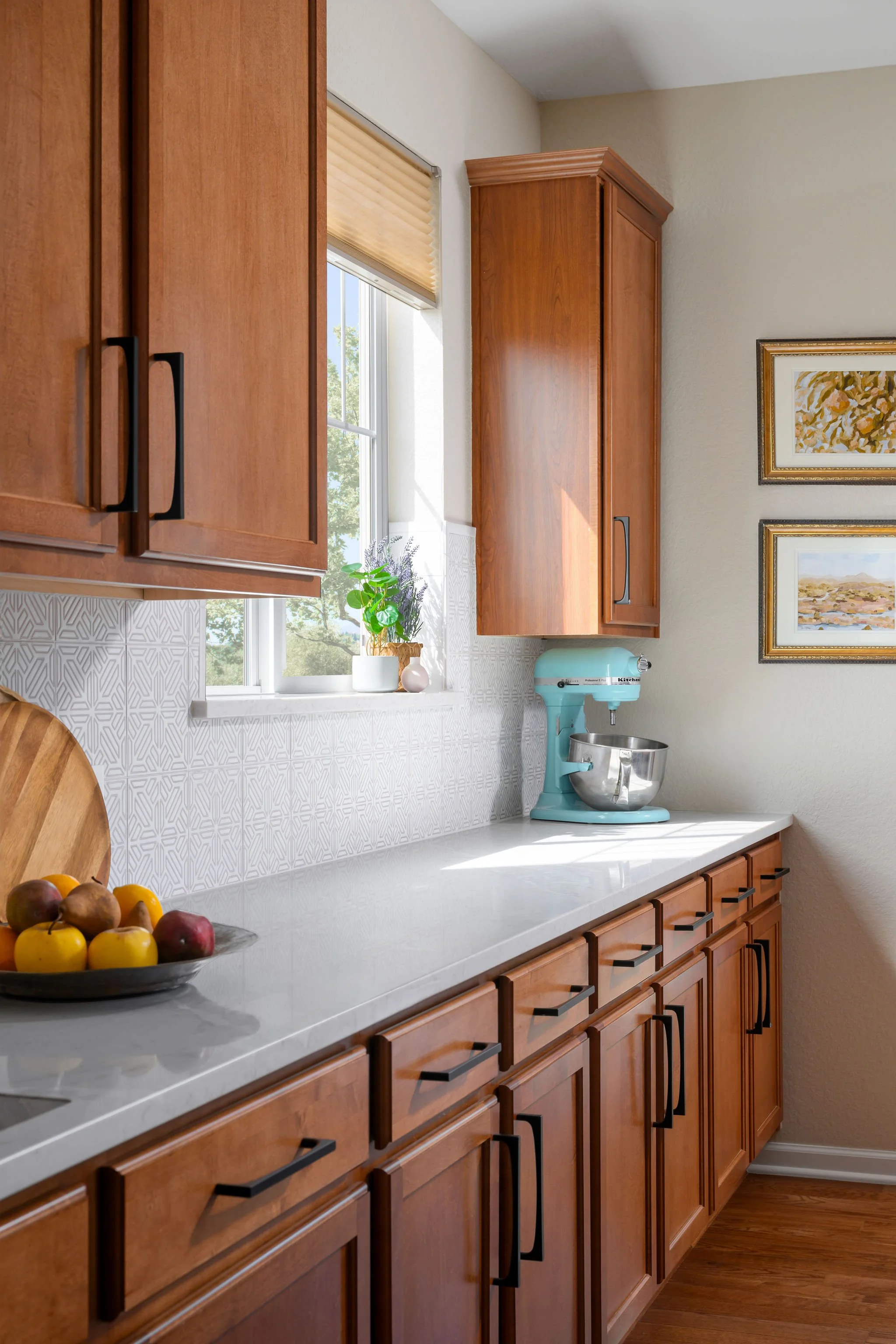 Kitchen with wooden cabinets, a window with blinds, a potted plant on the windowsill, a blue stand mixer on the countertop, a fruit bowl with apples and lemons, and framed landscape art on the wall.