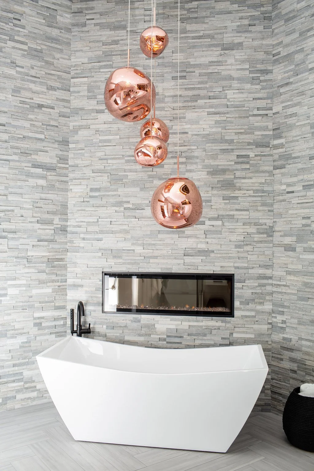 Modern bathroom with a white freestanding bathtub, a black floor-mounted faucet, a rectangular fireplace set into a textured gray stone wall, and a set of five large copper pendant lights hanging from the ceiling.