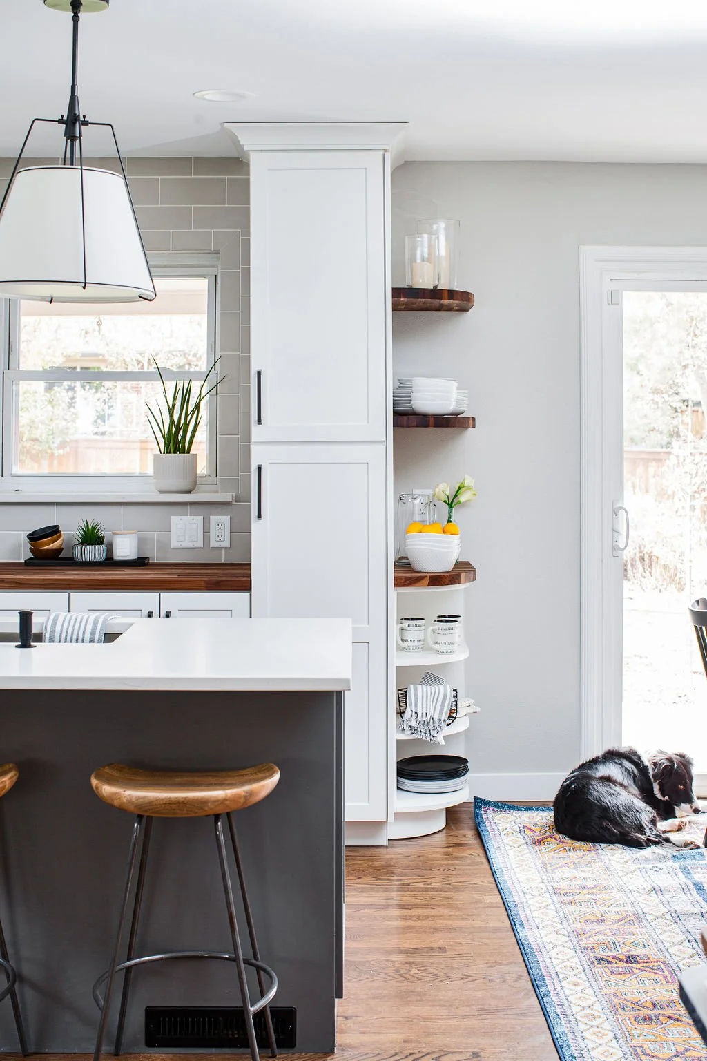 Modern kitchen interior with white cabinets, wooden countertops, open shelving, a window with a potted plant, and a black and white dog lying on a colorful rug near a sliding glass door.
