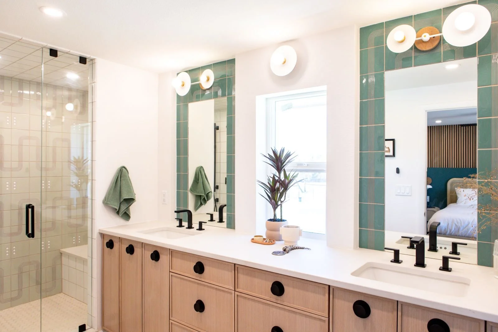Modern bathroom with double vanity, black faucets, large mirrors framed with green tiles, glass shower enclosure, potted plant, and green towels.
