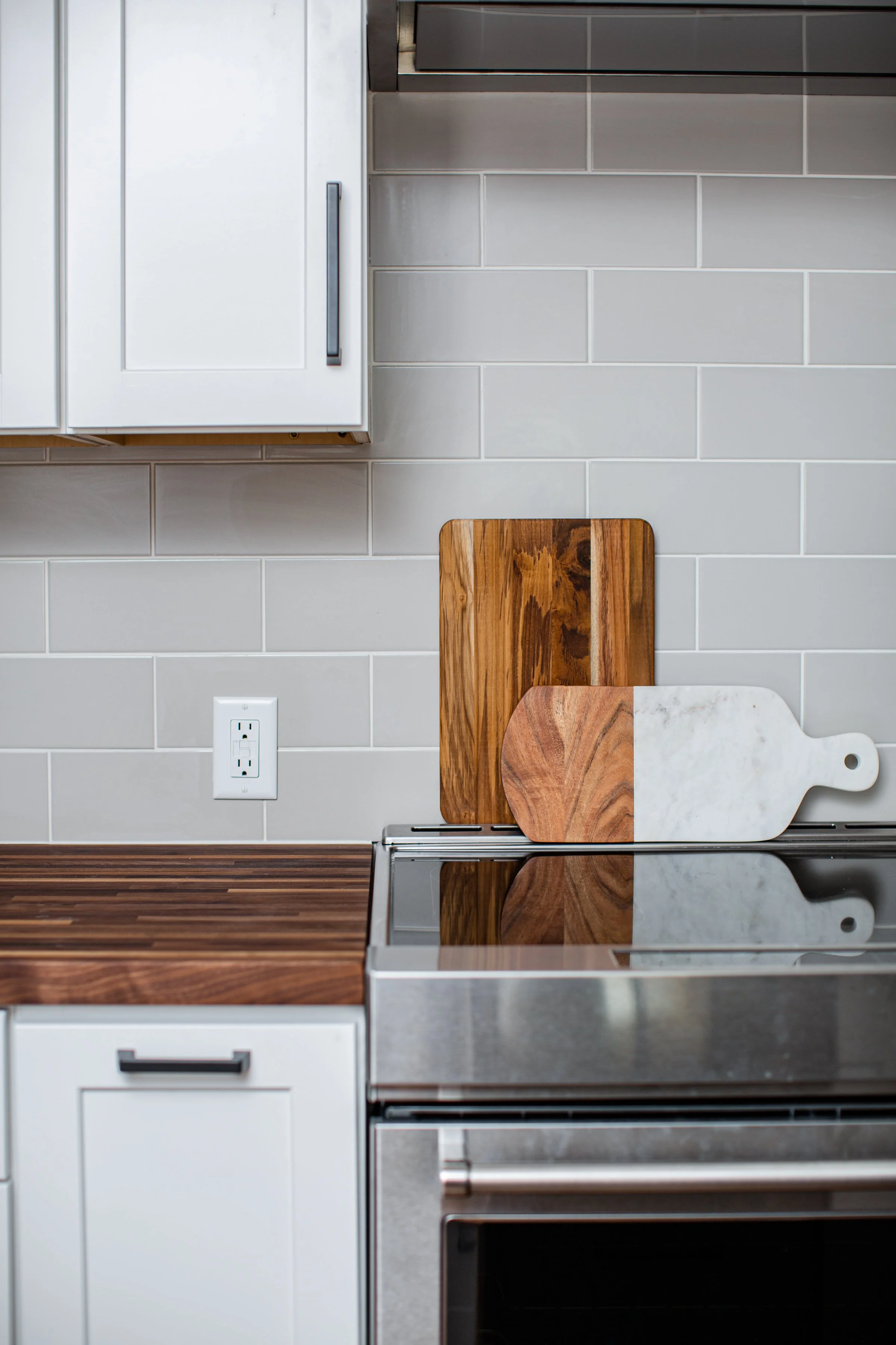 Kitchen countertop with two wooden cutting boards, single white marble cutting board, electric stove, completed in modern style with white cabinetry, gray backsplash tiles, and an electrical outlet.