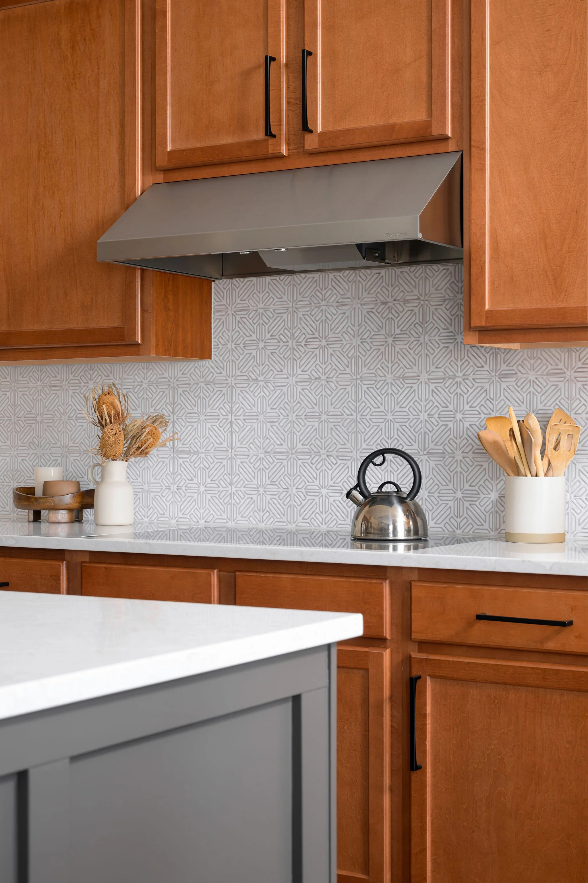 Kitchen with wooden cabinets, a gray range hood, a patterned gray backsplash, a kettle on the countertop, and white containers with wooden utensils and dried flowers.
