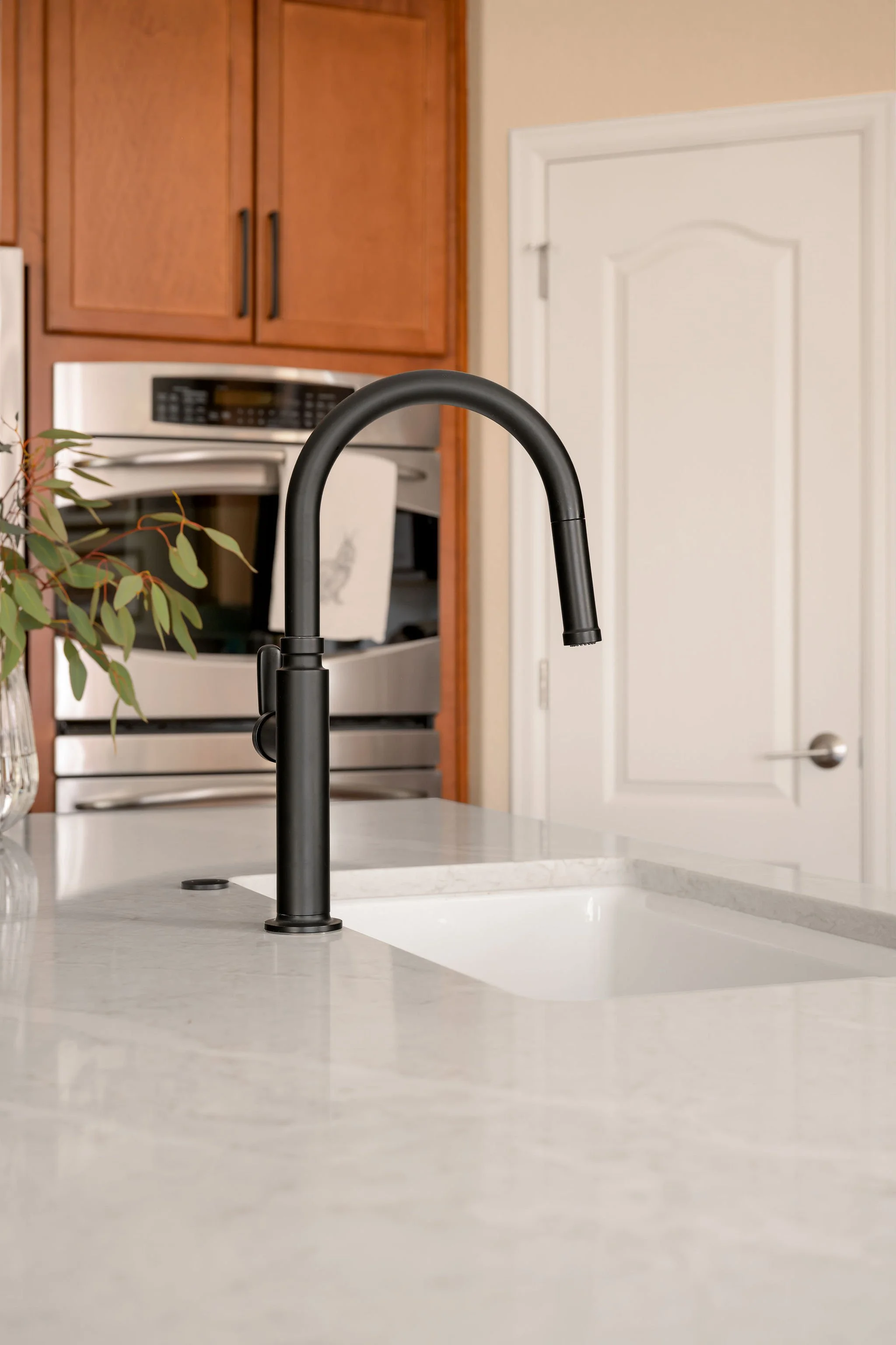 A black kitchen faucet mounted on a white marble countertop with a white inset sink, wooden cabinets, and a stainless steel oven, in a kitchen with light-colored walls.