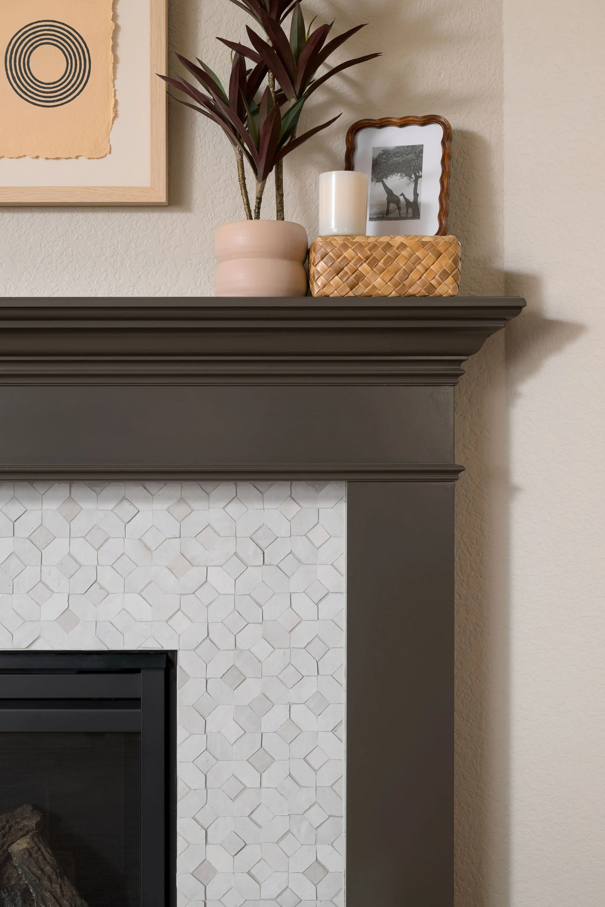 Close-up of a fireplace mantel decorated with a potted plant, a candle, a framed picture of a giraffe, and a woven box, with part of a wall featuring textured beige paint and a piece of abstract wall art.