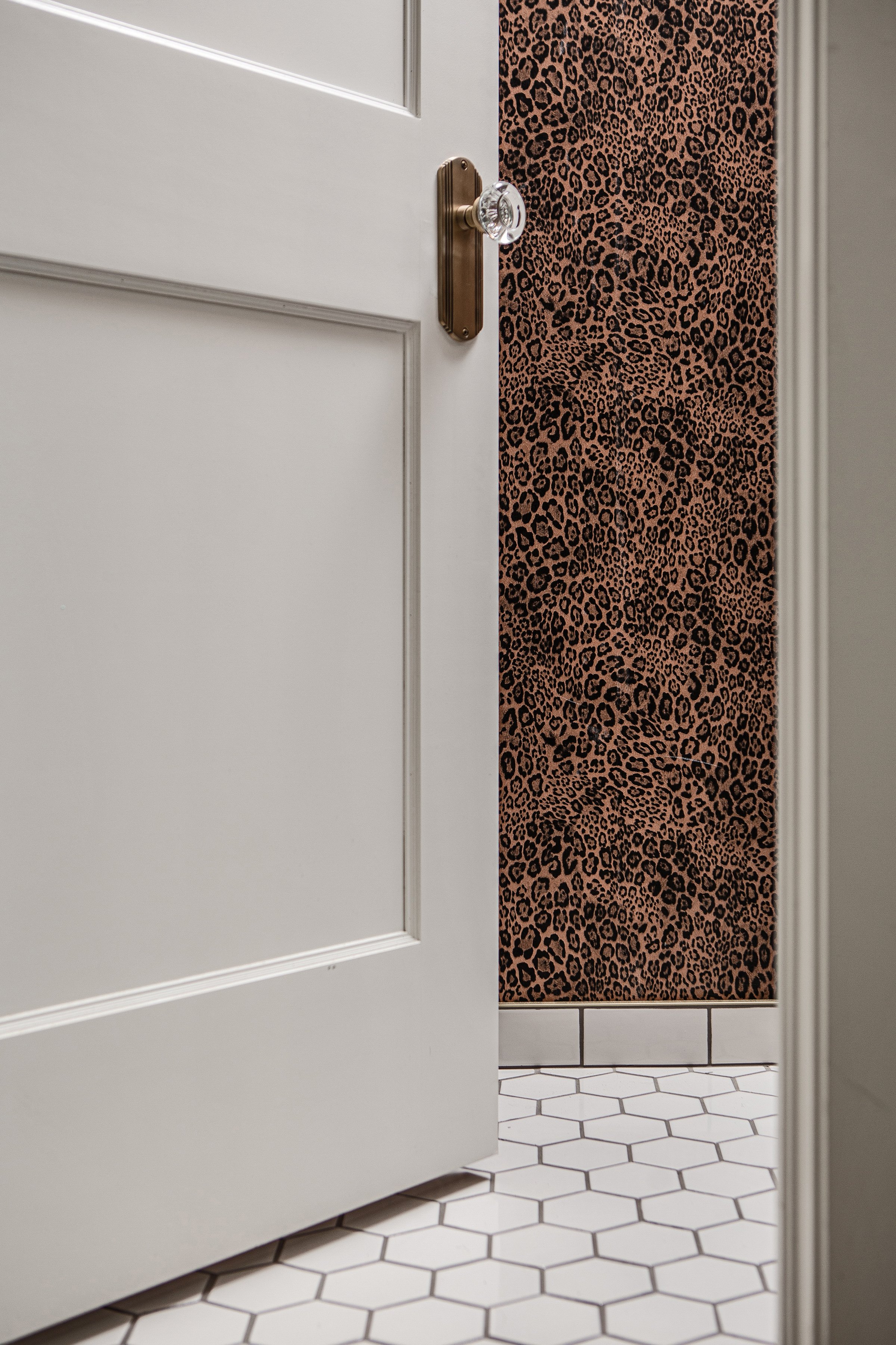 Partially open white door with a door knob, revealing a leopard print wall and white hexagonal tile flooring inside a room.