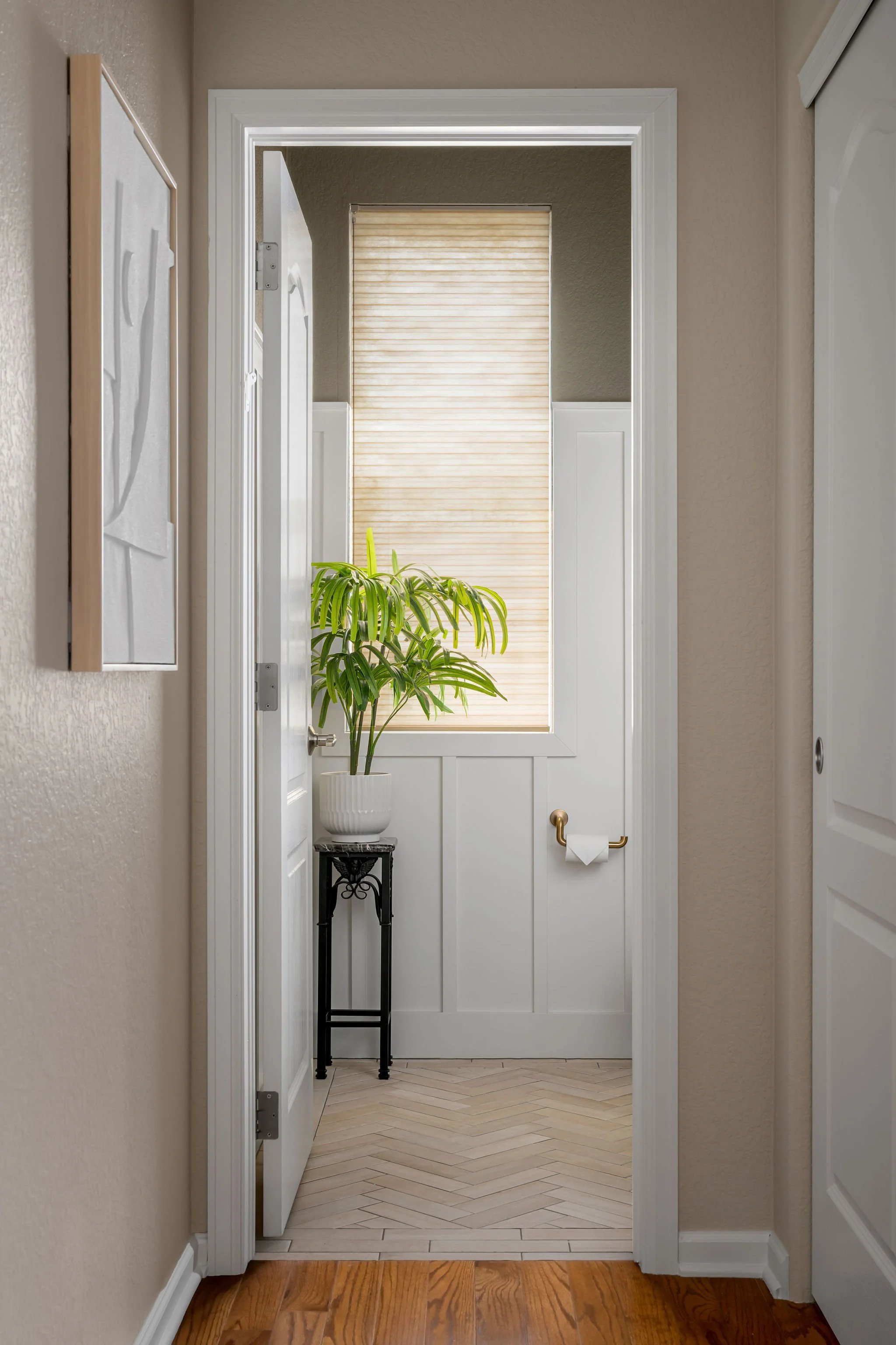 A small interior room with a potted plant on a black stand near a window with a beige blind, white wainscoting, beige walls, and a wooden floor.
