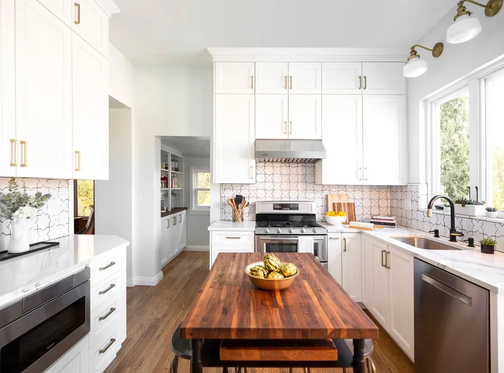 A bright kitchen with white cabinets, a wooden dining table, stainless steel appliances, and a window letting in natural light.