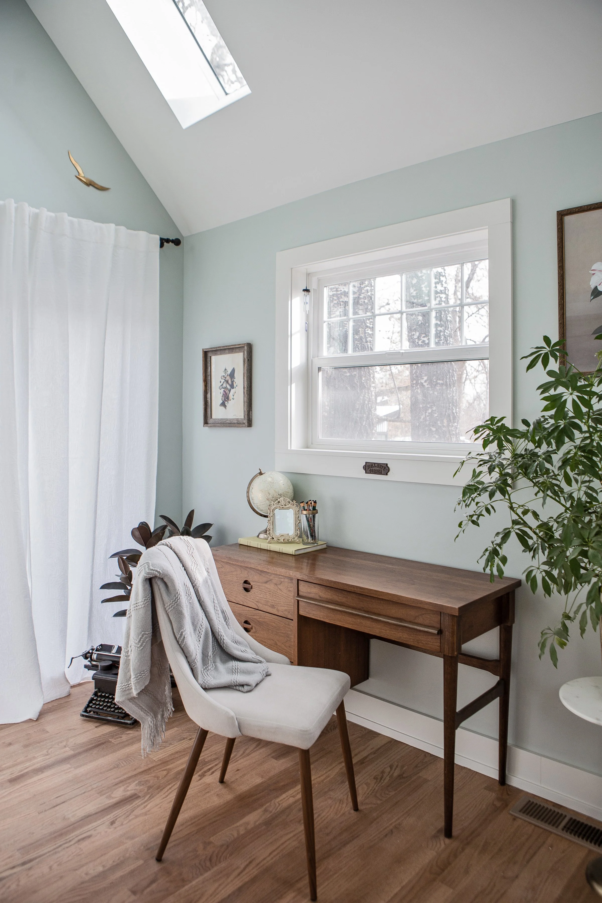 A bright corner of a room with a wooden desk, a white upholstered chair with a gray blanket, a small globe, and a mirror on the desk. There is a window and a white curtain, with framed artwork and a large houseplant nearby.