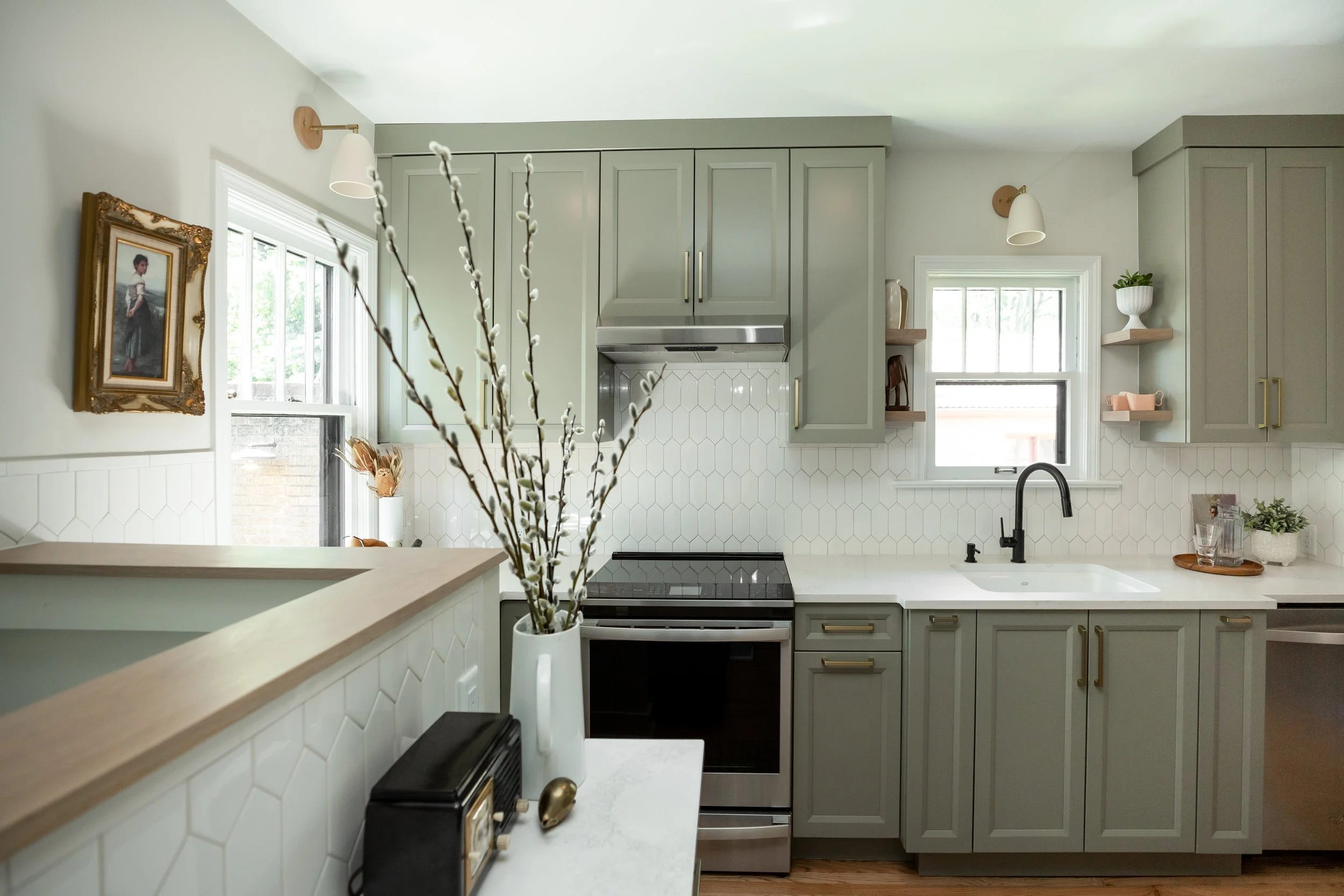 Modern kitchen with light green cabinets, white hexagonal tile backsplash, black faucet, and small decorative shelves, decorated with a vase of pussy willow branches and small art pieces.
