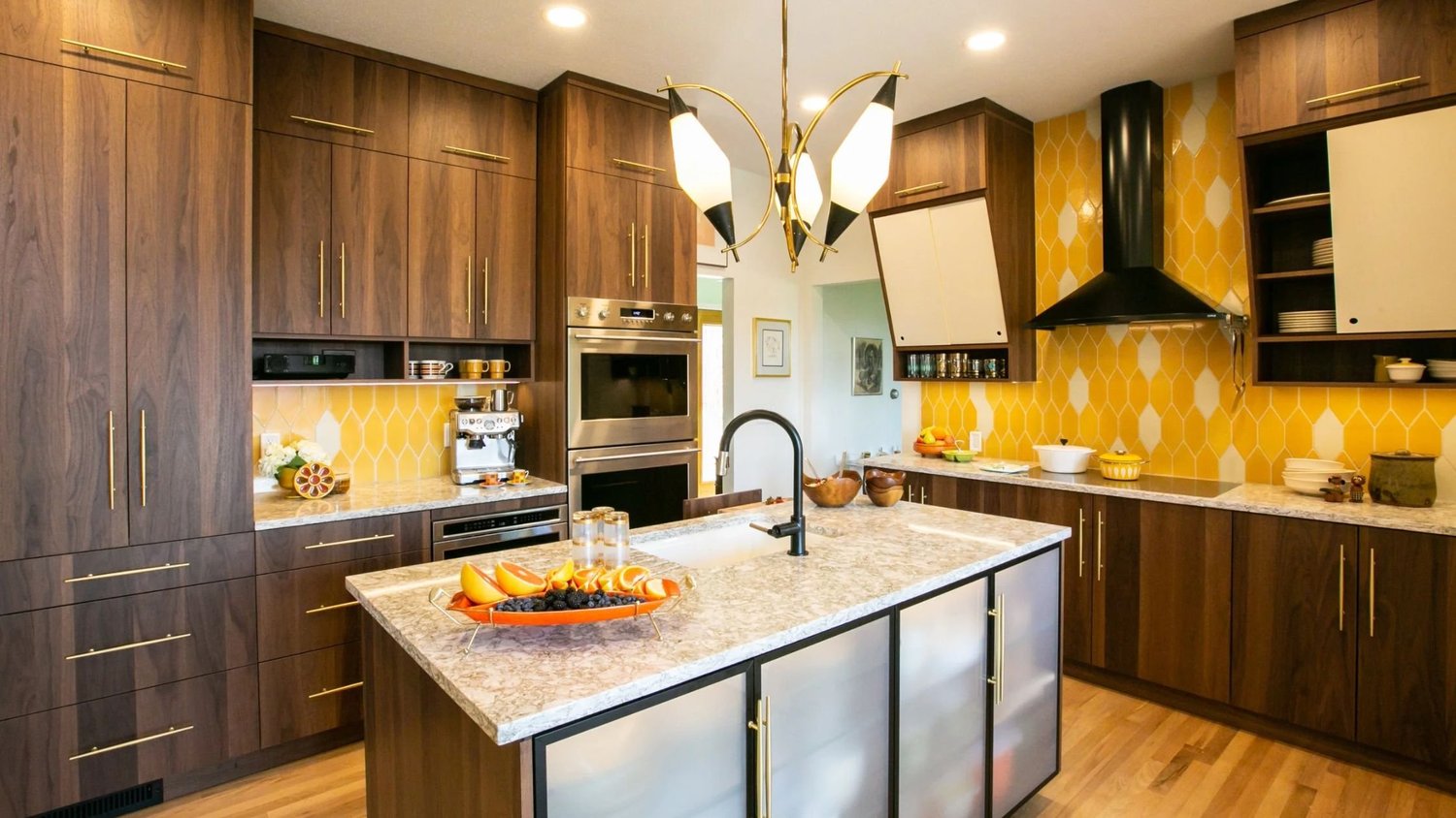 Mid-century modern kitchen with walnut cabinetry, a yellow hexagon tile backsplash, brass hardware, a black range hood, and an island with a speckled stone countertop