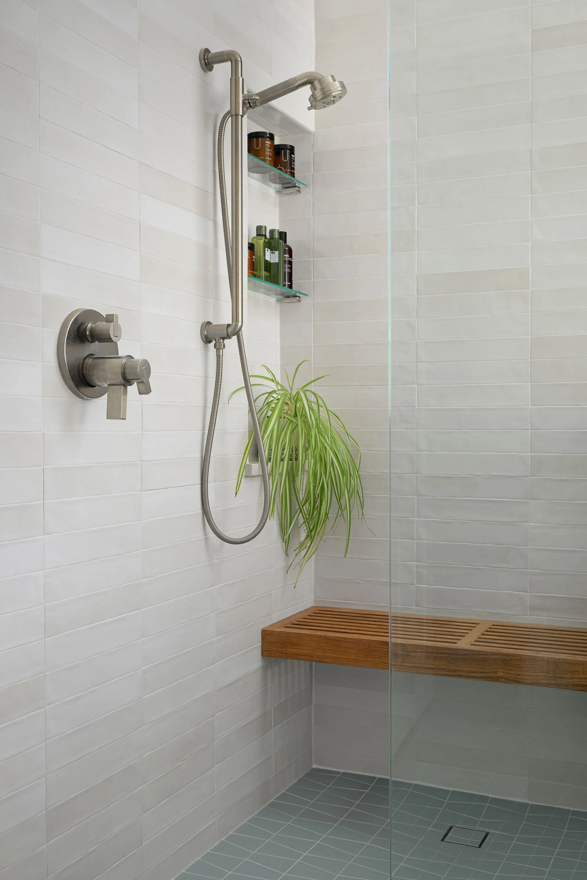 A modern shower with white tiled walls, a wooden bench, built-in glass shelves with bottles, a green potted plant, and a ceiling-mounted showerhead.