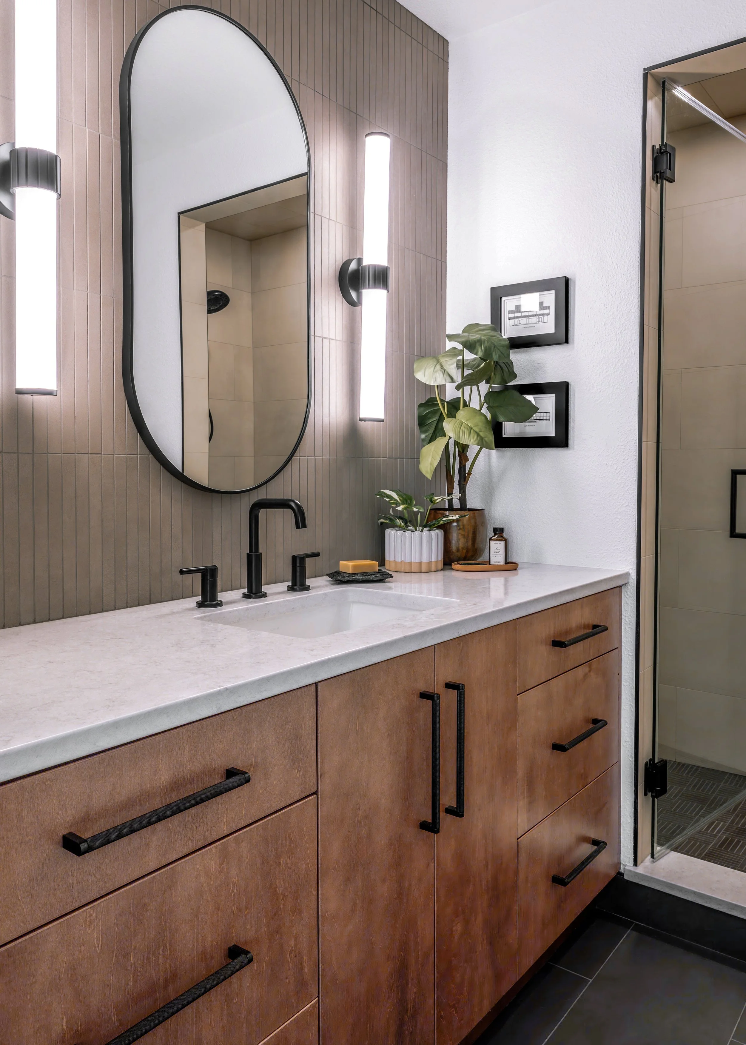 Modern bathroom vanity with a white countertop, black fixtures, and wooden cabinets. An oval mirror and vertical light fixtures are mounted on a textured brown tile wall. A potted plant and framed artwork are on the countertop, and a glass shower door is visible to the right.