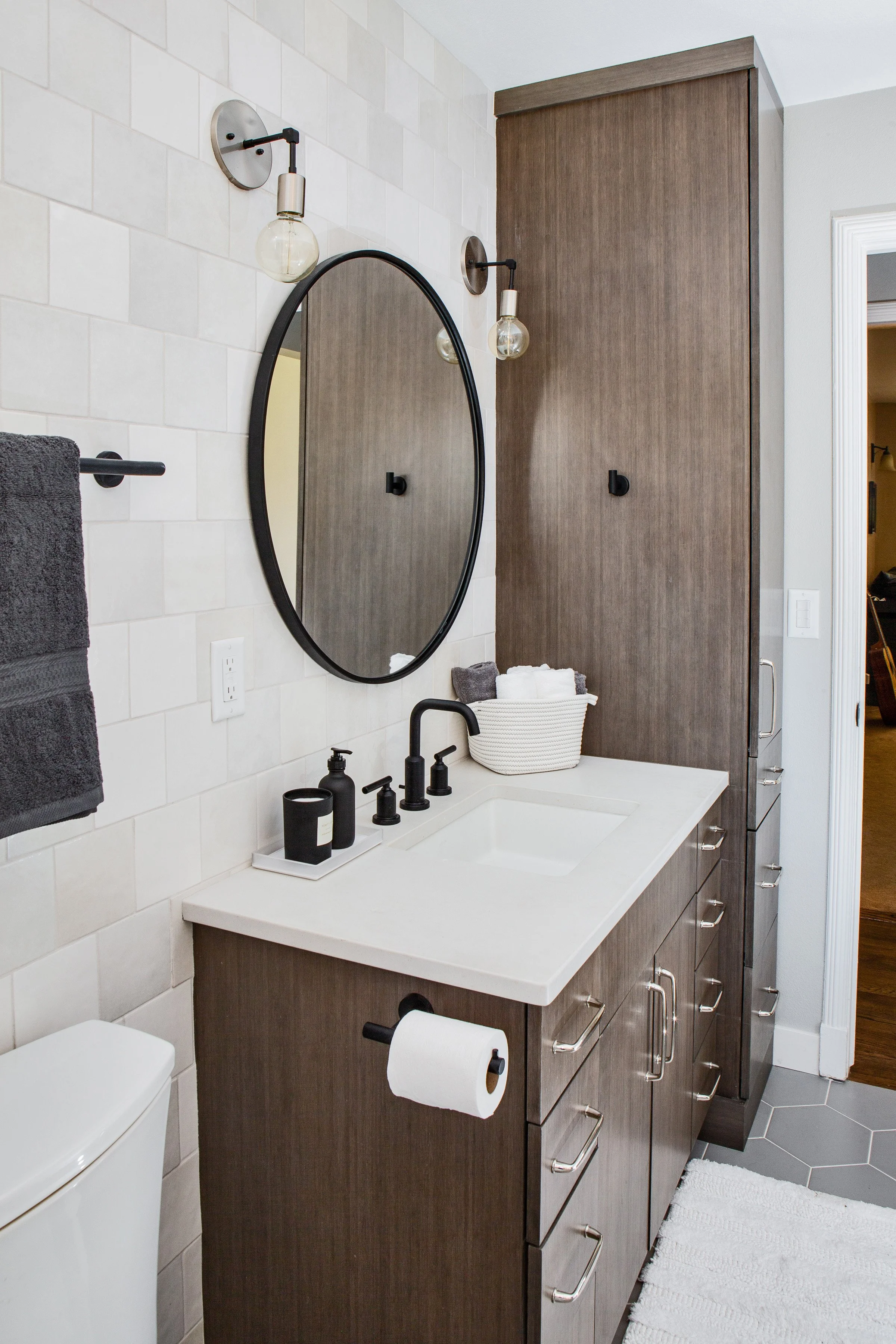 Modern bathroom vanity with a white countertop, black fixtures, an oval mirror, and a tall wooden cabinet, with black wall-mounted lights and a towel hanging on a black bar.