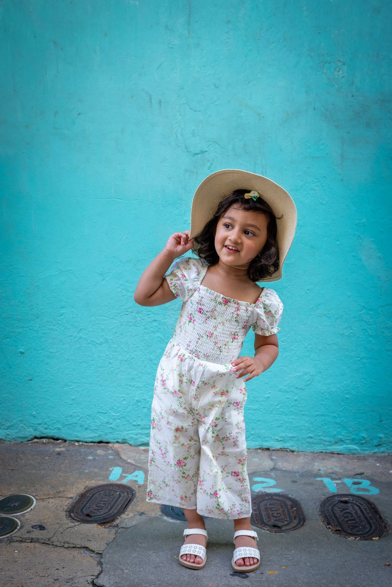 A young girl wearing a floral dress and white sandals, holding a wide-brimmed straw hat, standing against a bright blue wall, smiling and touching her hair.