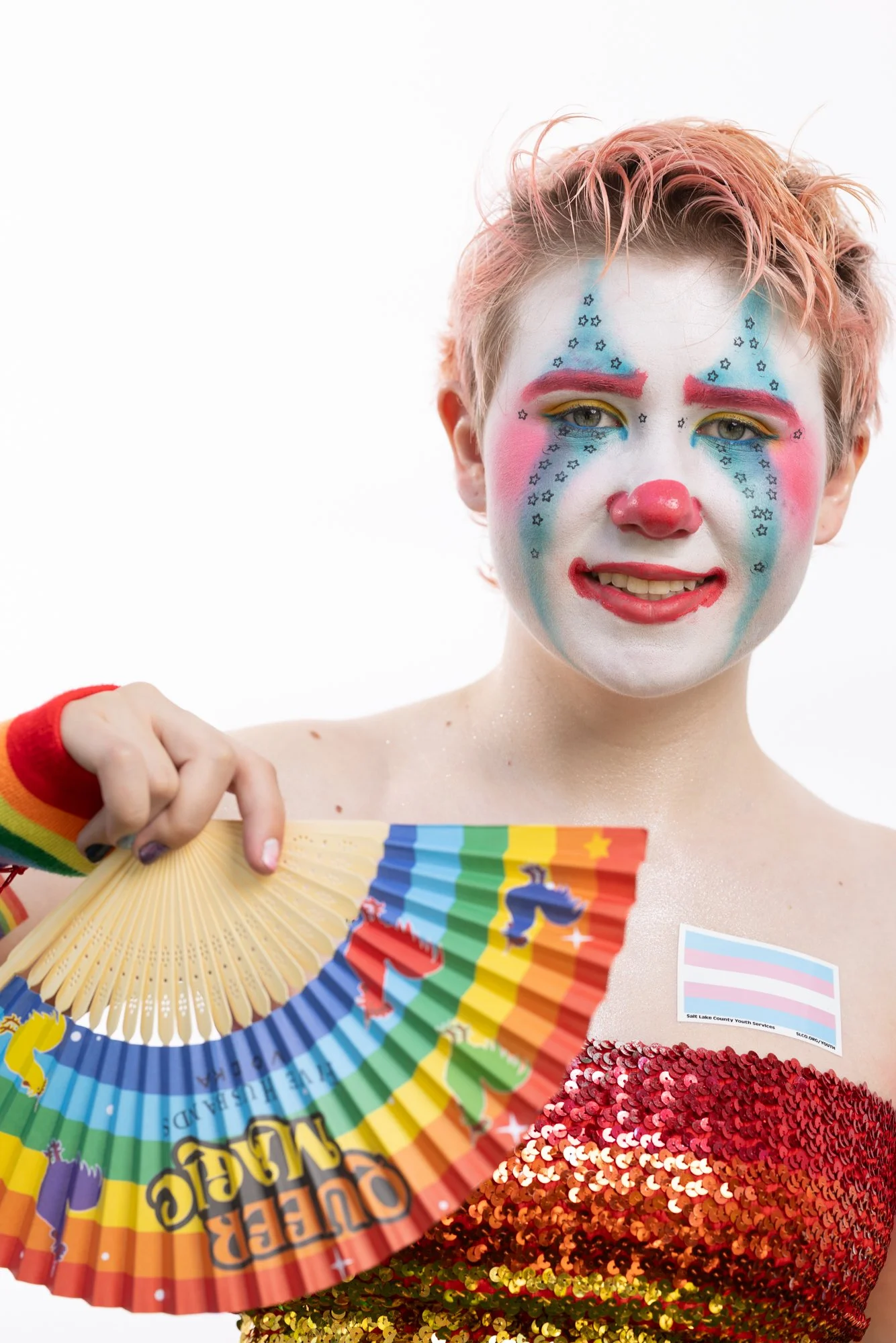 Person with colorful clown makeup, including white face paint, red and blue accents, and star patterns, holds a rainbow Pride fan and wears a red sequined top and rainbow wristbands.