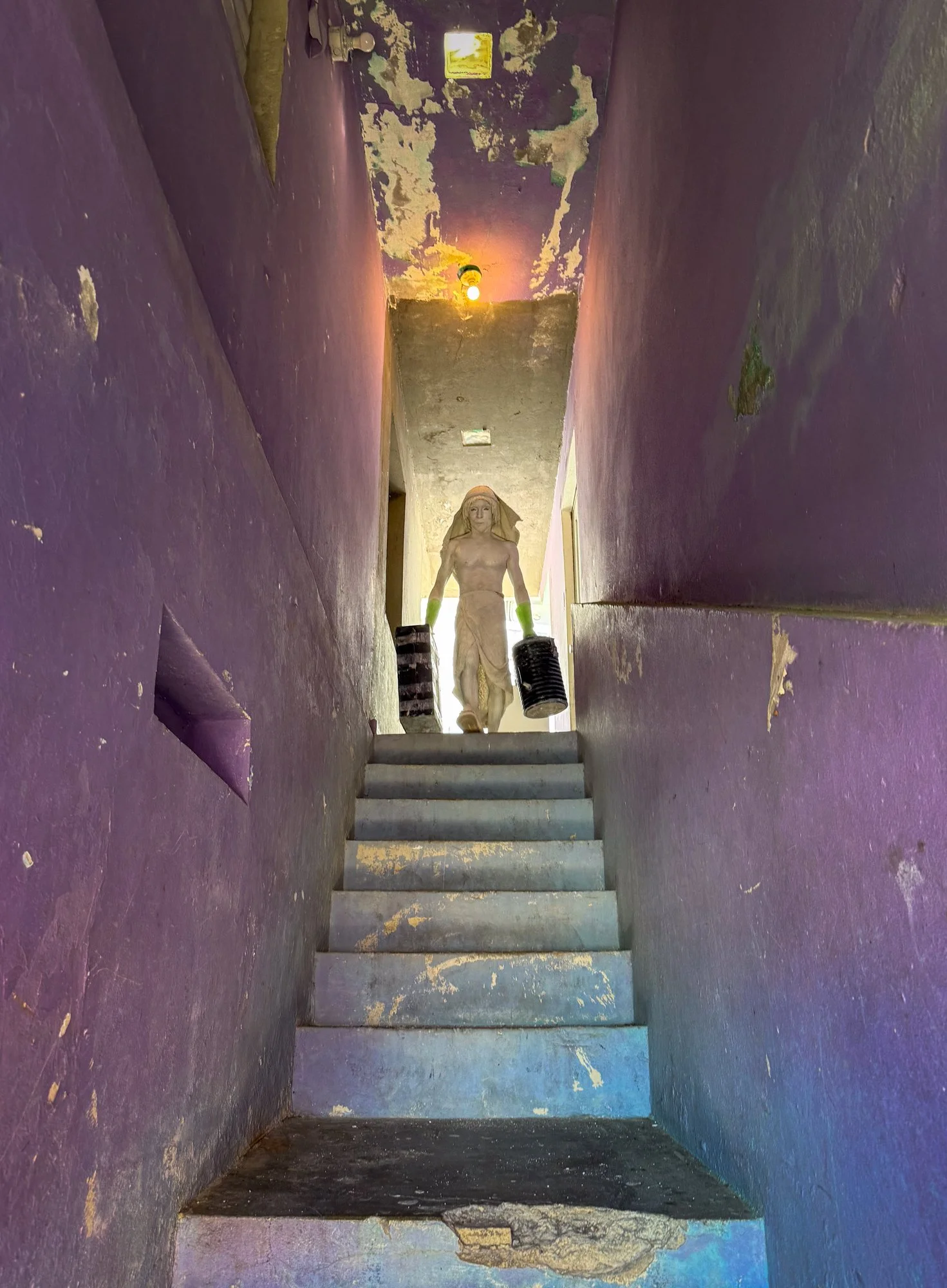 A set of worn stairs leading to a small landing with a humanoid statue holding two black buckets, in a narrow, purple-walled corridor with peeling paint and a ceiling light.