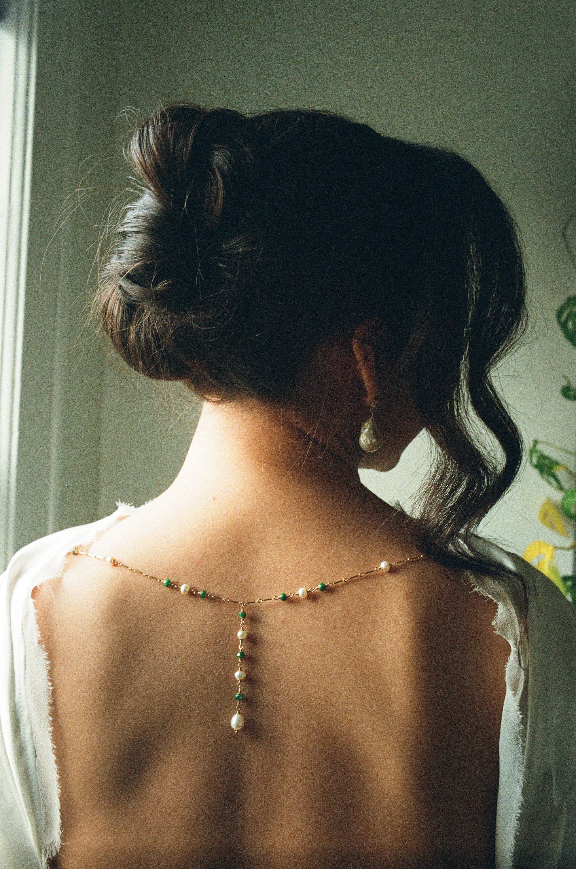A woman with dark, wavy hair styled in an elegant updo, wearing pearl and green beaded jewelry, is facing away from the camera, revealing her shoulder and neck, with a softly lit background.