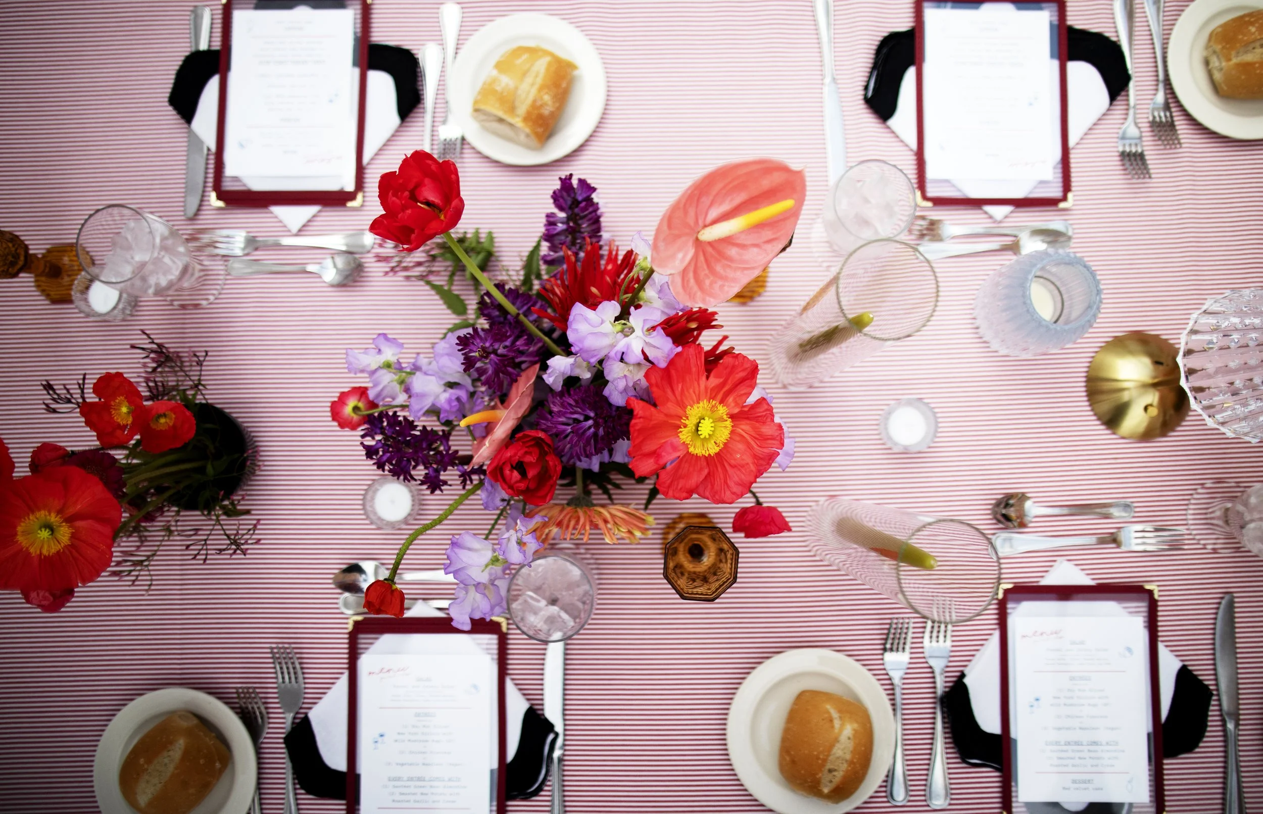 A top-down view of a formal dining table with a red and white striped tablecloth, featuring a large colorful flower centerpiece, place settings with menus, bread rolls, glasses, and cutlery.