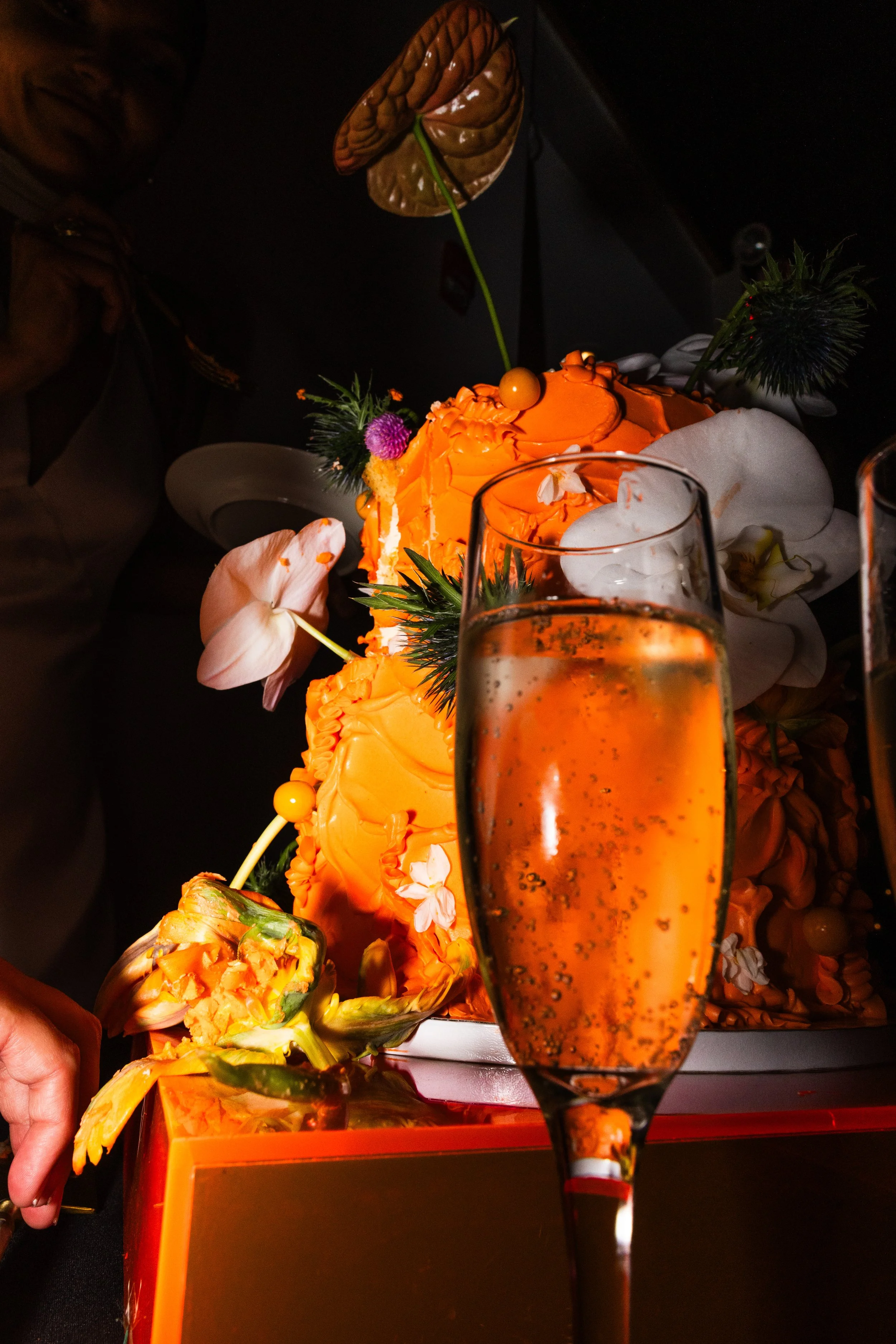 A slice of orange-colored cake with decorative flowers, greenery, and an artistic edible flower decoration, with a glass of sparkling drink in front.