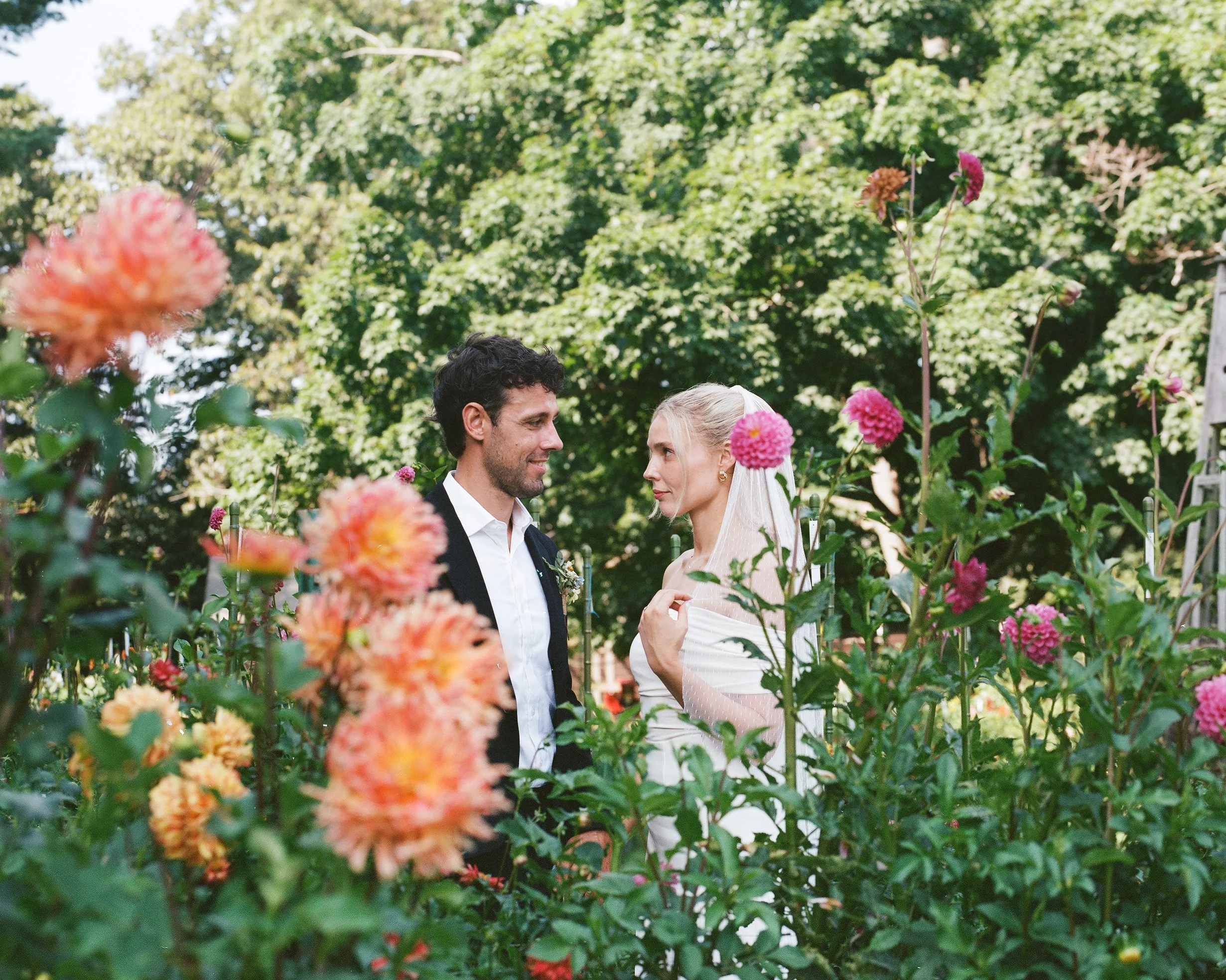 A couple dressed in wedding attire standing closely together in a lush garden surrounded by pink and orange flowers, with green trees in the background.