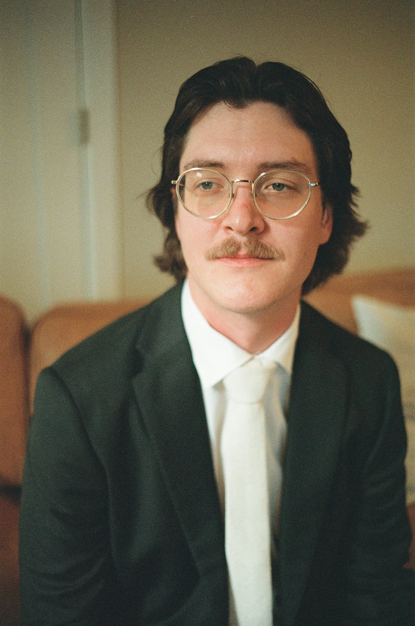 Man with dark hair, glasses, and mustache, wearing a tuxedo with a white shirt and light-colored tie, sitting in a room.
