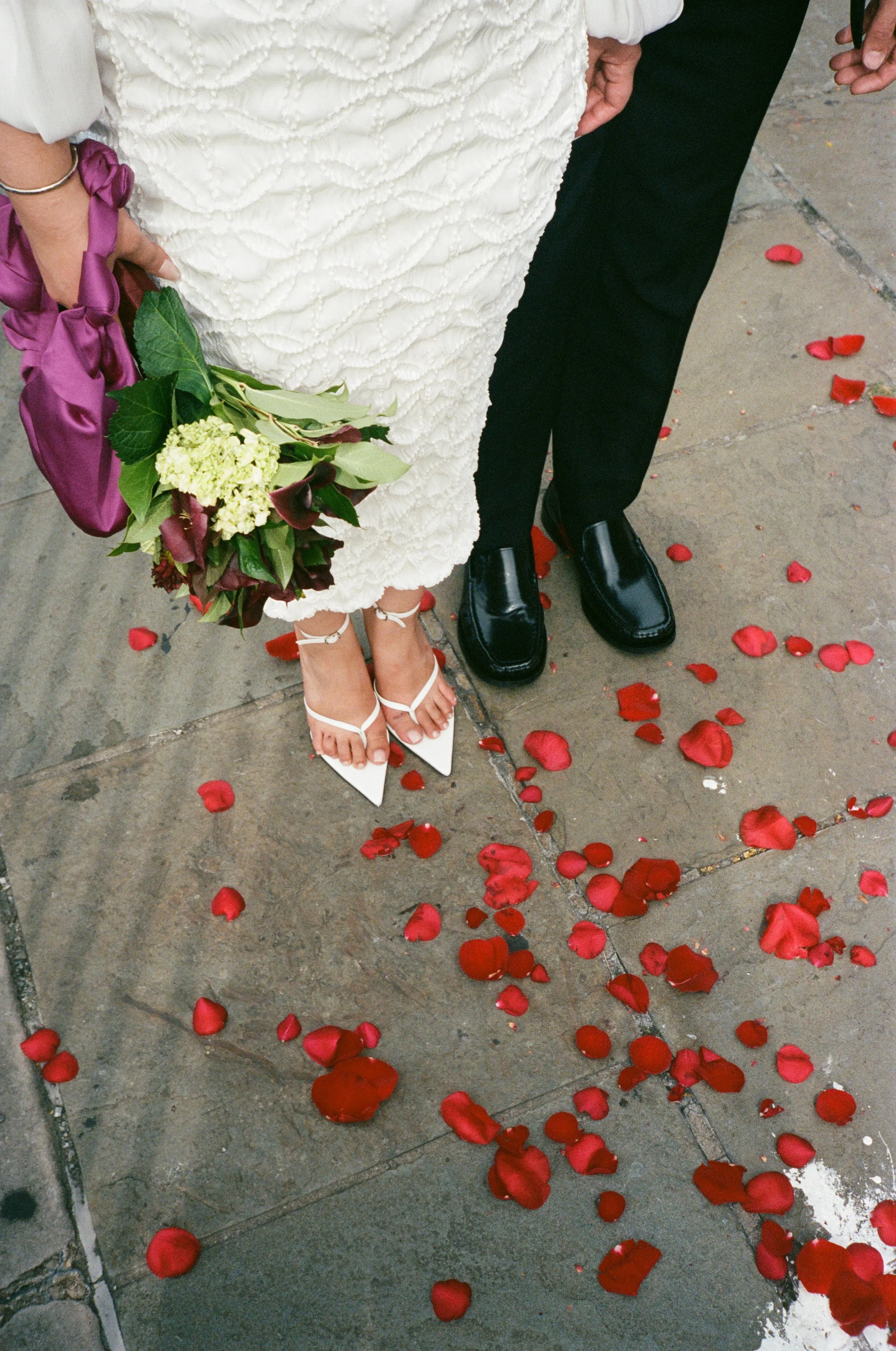 Close-up of two people standing on a sidewalk with scattered red rose petals. One person is wearing a white textured dress and white strappy heels, holding a bouquet of green and purple flowers. The other person is dressed in black shoes and trousers