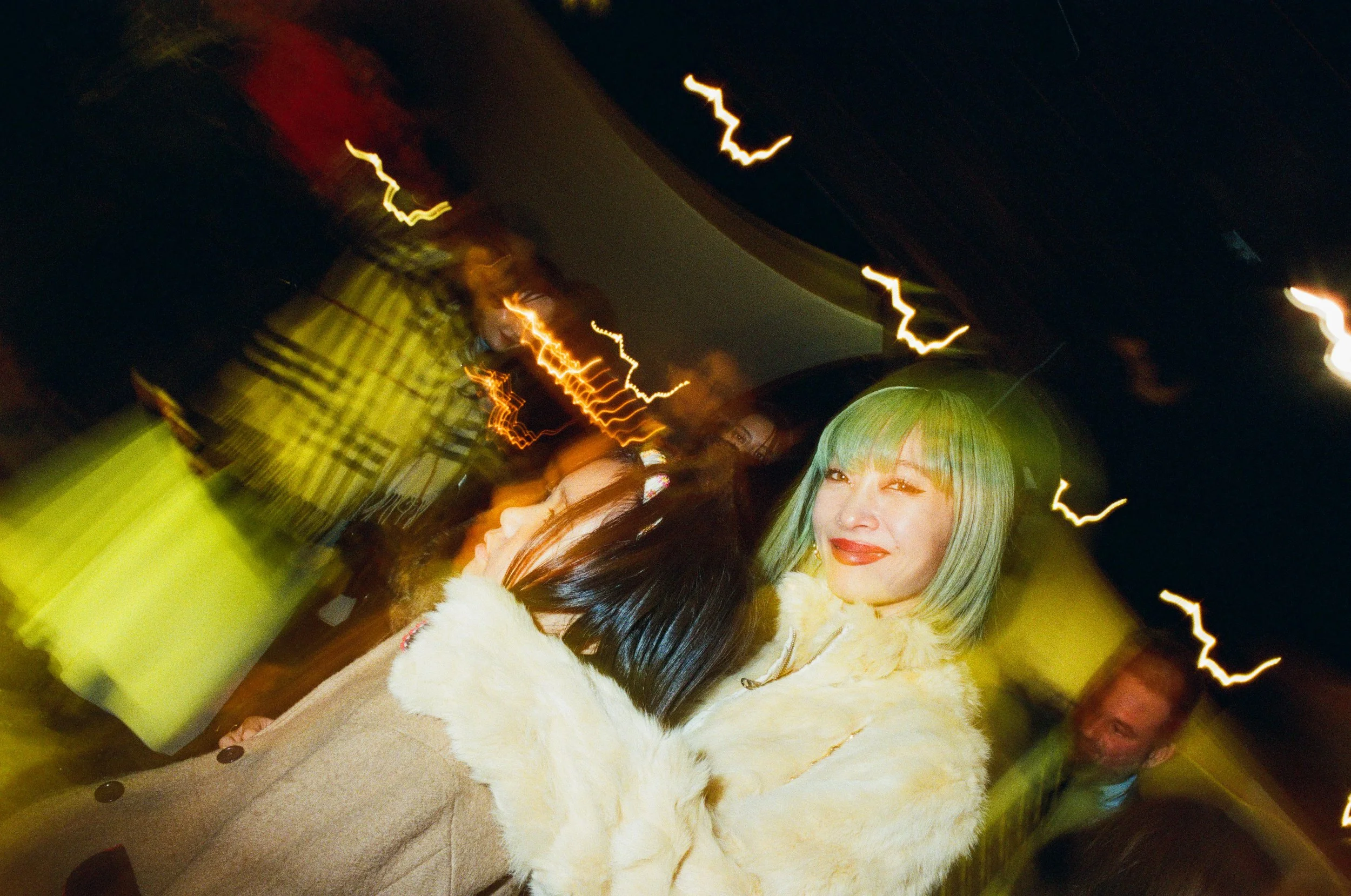 Two women and a man at a nighttime event with bright yellow lighting and light streaks in the background. One woman with colored hair is smiling, while the other woman with dark hair is partially visible. The man is in the background, smiling.