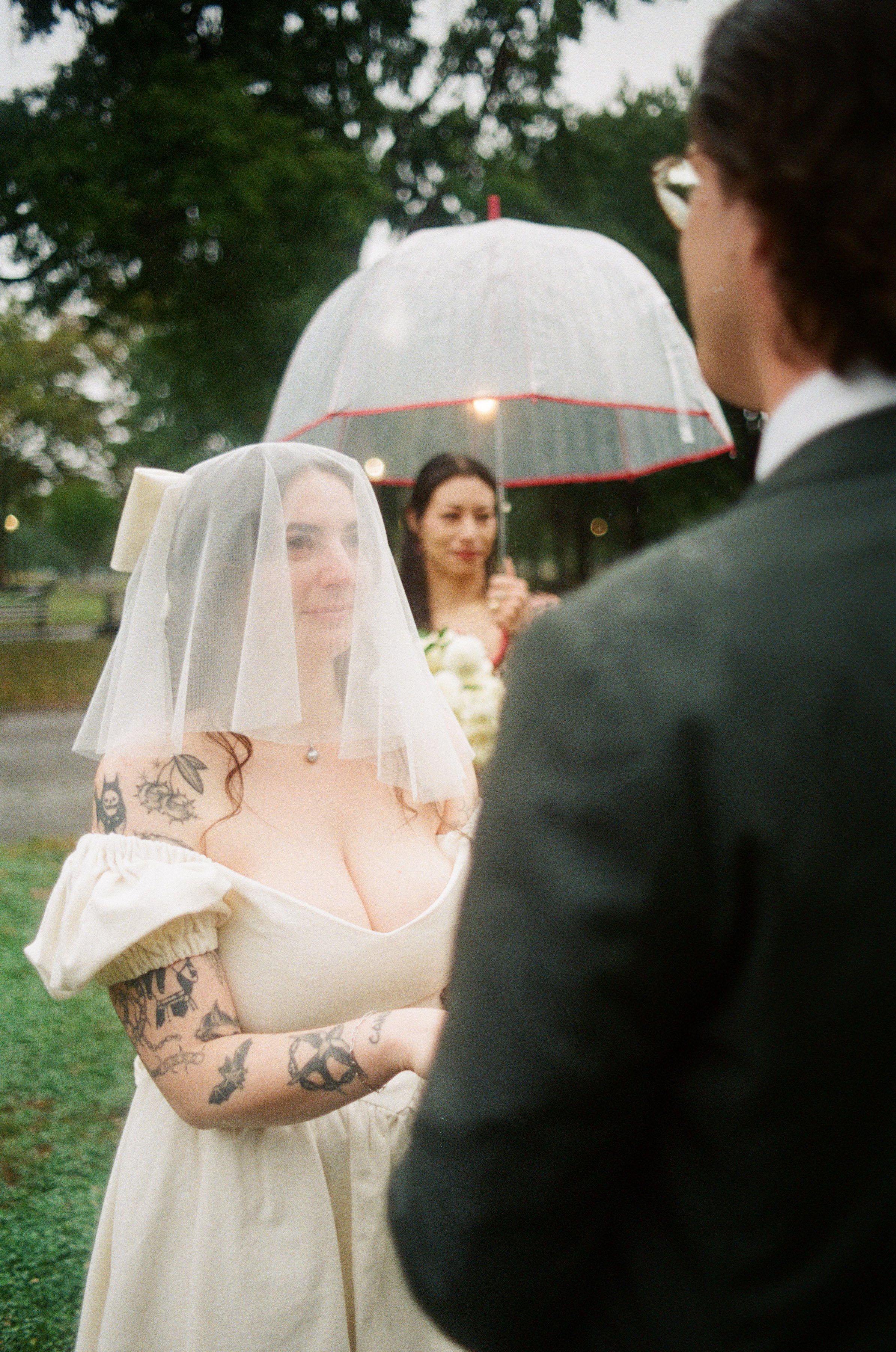 A bride wearing a white dress and veil with tattoos on her arms, standing outdoors during a rainy wedding ceremony, with a person holding a transparent umbrella in the background.