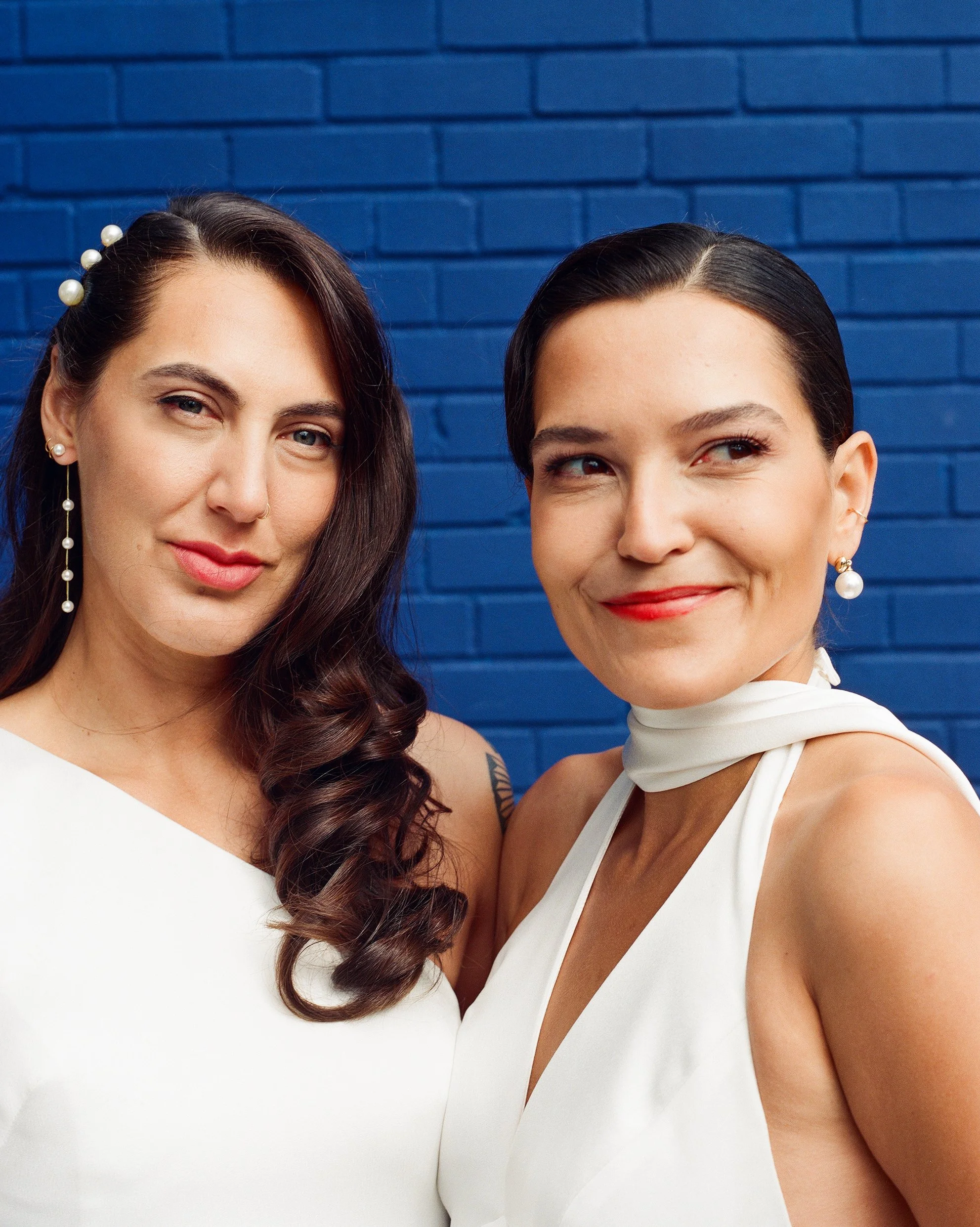 Two women smiling in front of a blue brick wall, wearing white dresses and pearl earrings.
