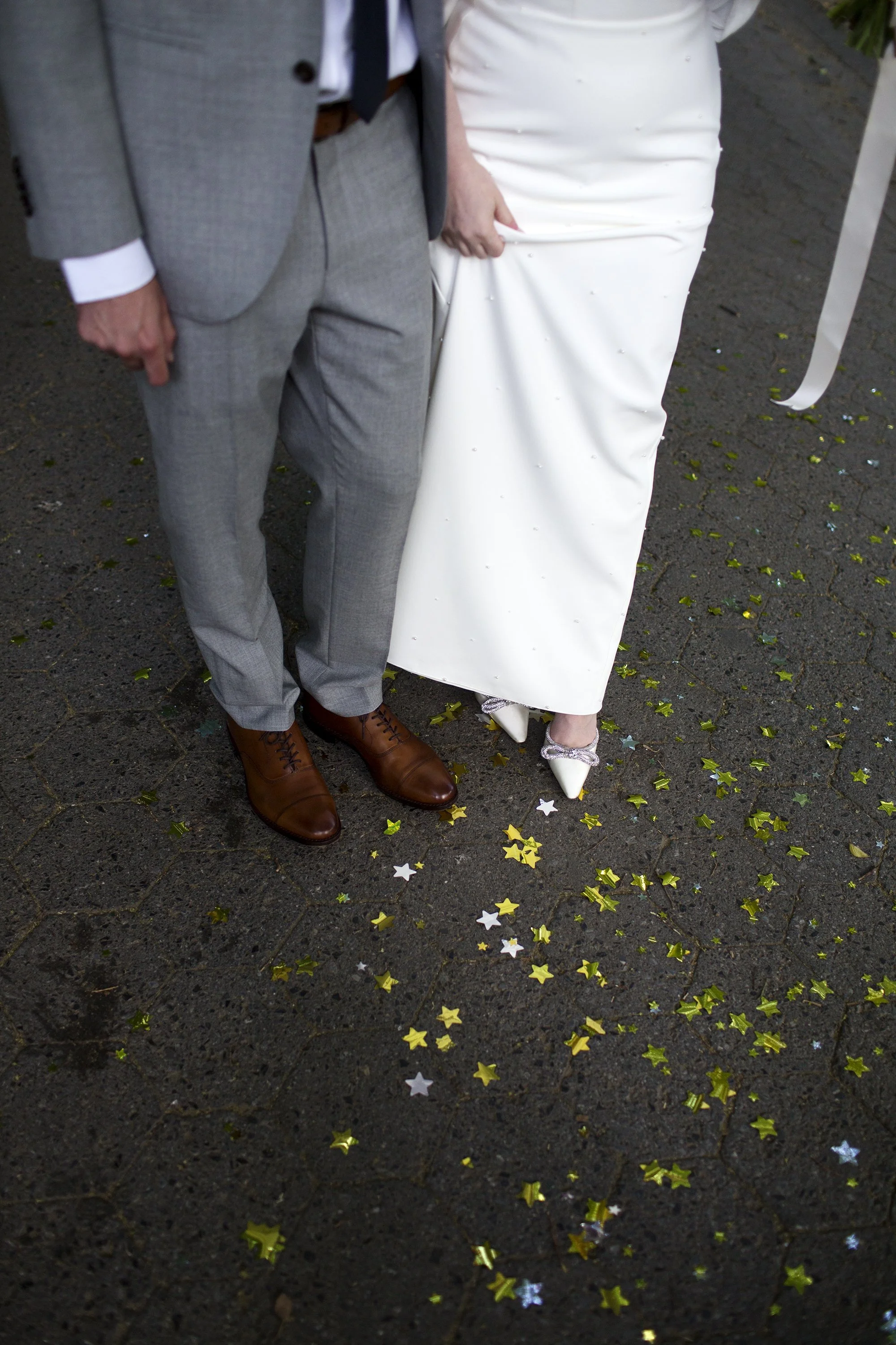 Close-up of a bride and groom at their wedding, showing only their lower bodies, standing on a dark pavement decorated with gold and silver star-shaped confetti.