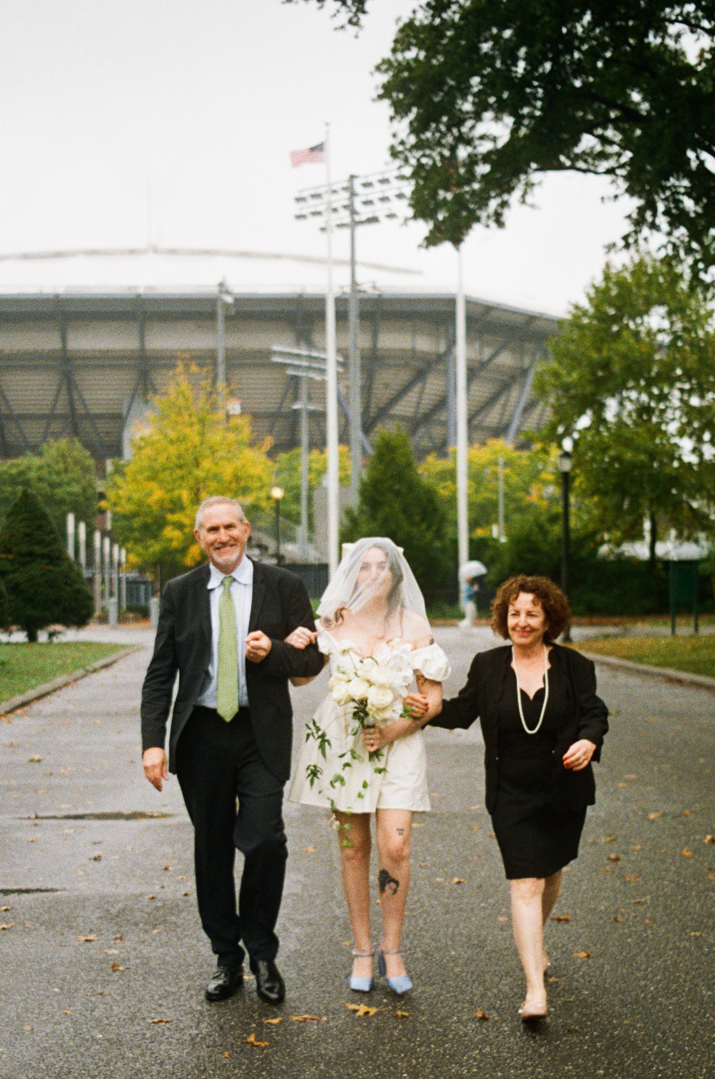 A woman in a wedding dress and veil walking between two older adults on a paved path in a park, with stadium lights and a large stadium in the background.