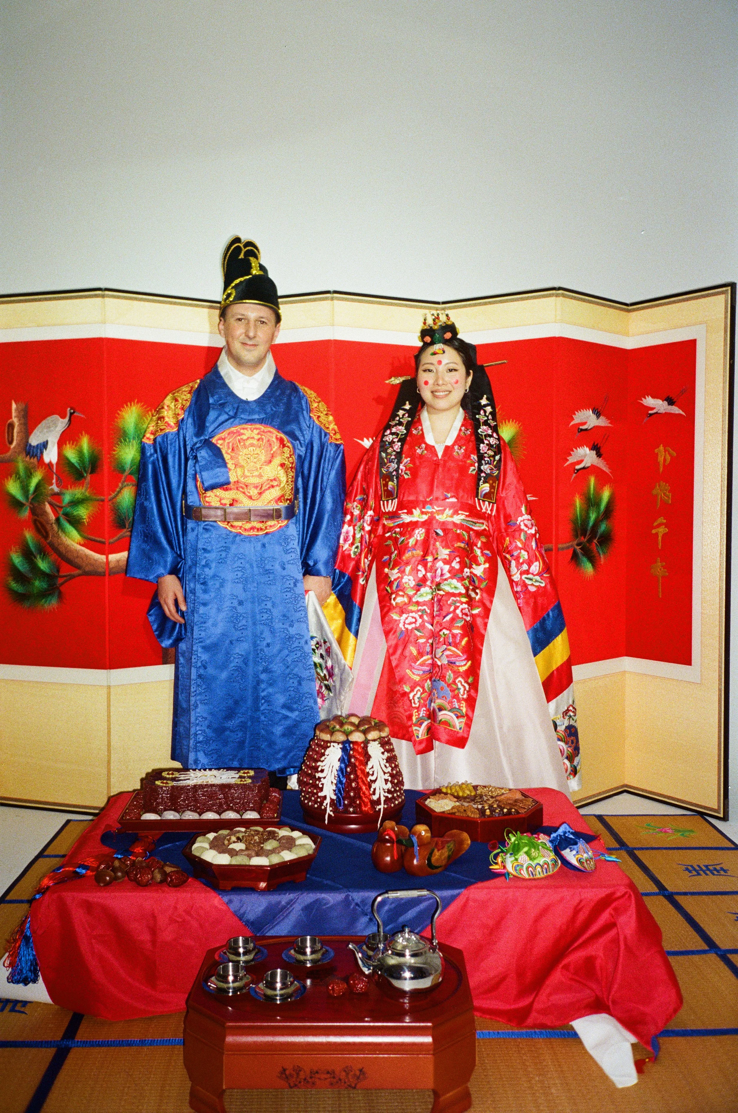 Two people in traditional Korean wedding attire standing behind a table with various traditional foods and decorations, with a red backdrop featuring cranes and Korean characters.